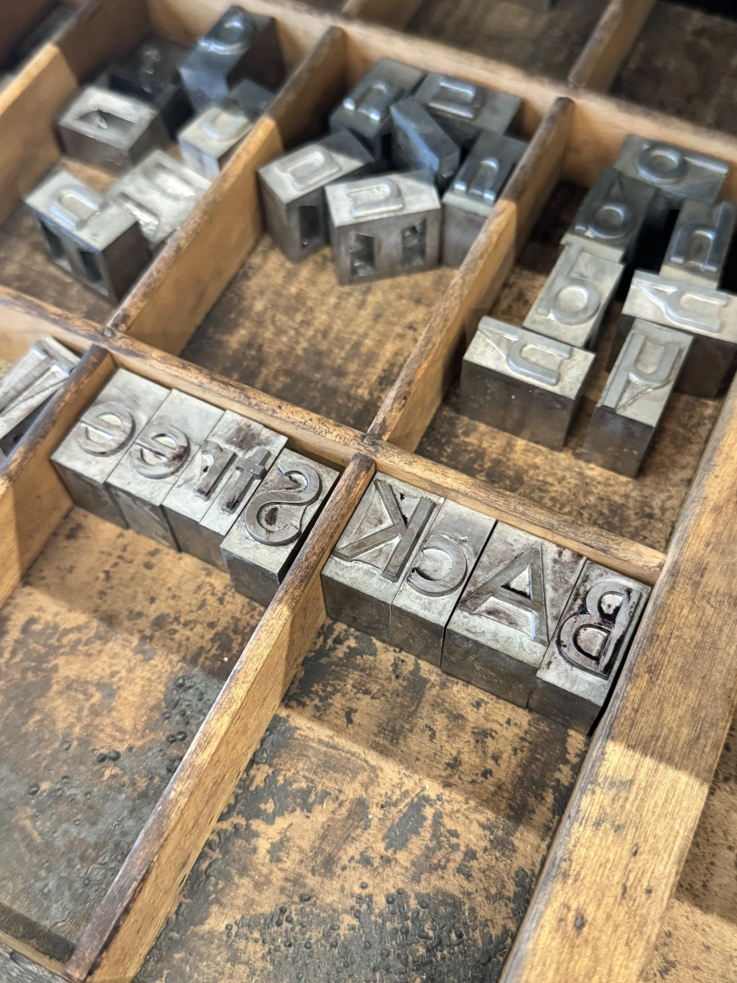 A wooden box with several metal letterpress type blocks inside, some showing letters such as 'D', 'E', 'A', 'Y', and 'N'.