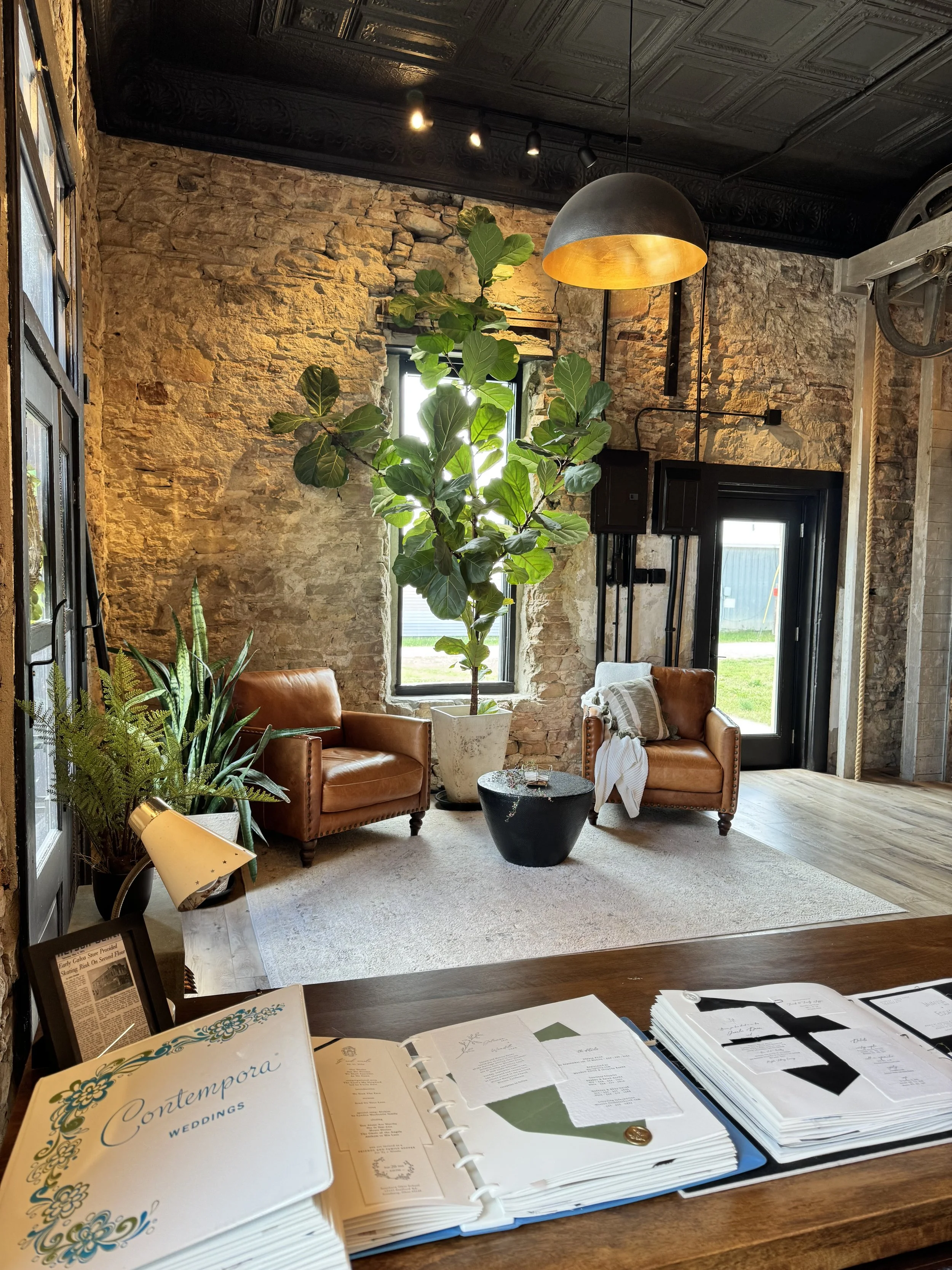 Indoor space with brick wall, large green plant, sitting area with two leather chairs, a window and glass door, hanging light fixture, and various printed materials on a table in the foreground.