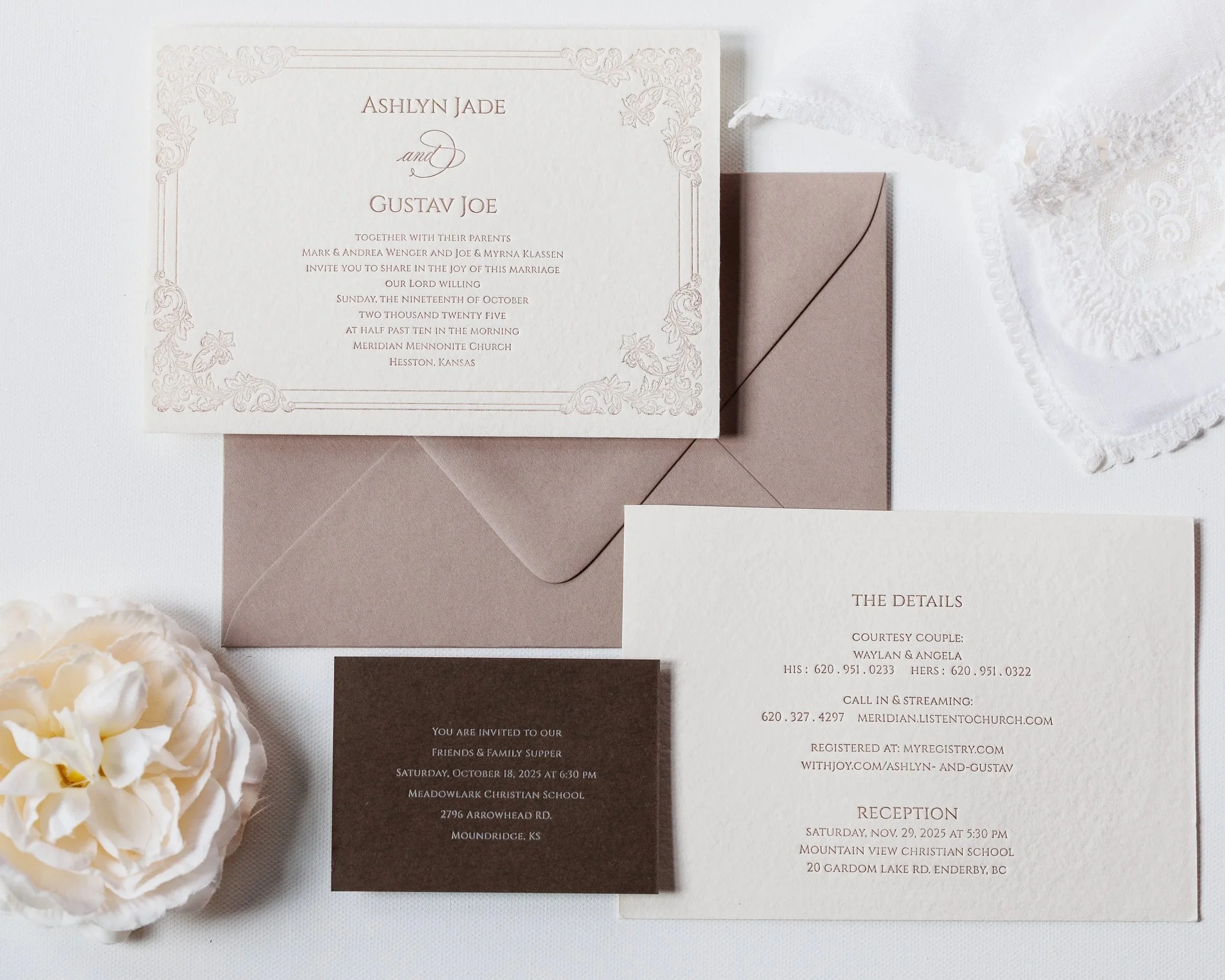 A wedding invitation card with intricate floral borders and elegant text announcing the wedding of Ashlyn Jade and Gustav Joe, along with event details and RSVP information, placed on a white textured surface with a brown envelope beneath it and white flowers to the side.