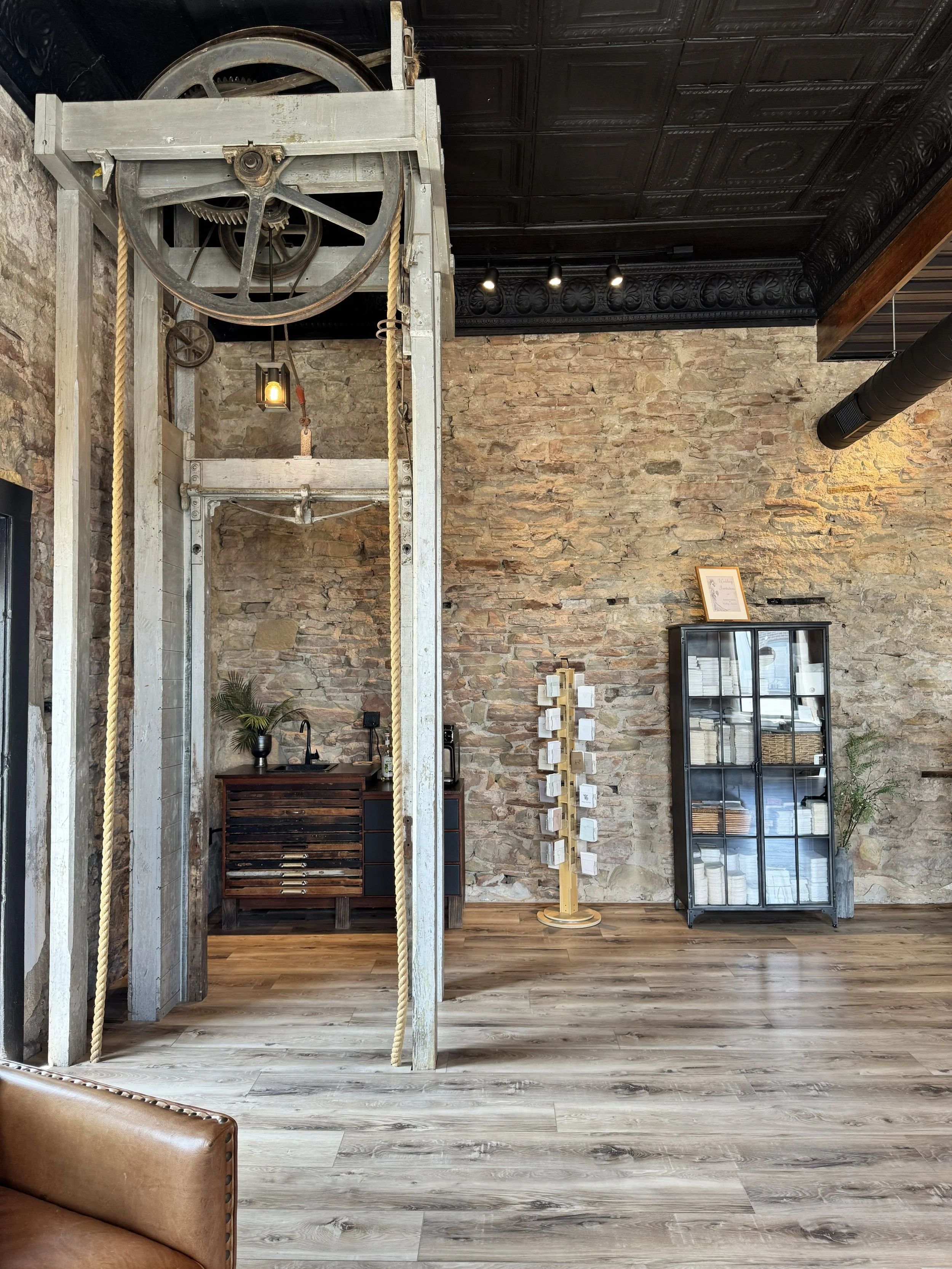 Interior of a modern, rustic-style room with exposed brick wall and wooden flooring. There is a black cabinet with glass doors, a small framed picture on top, a plant in a pot, and a wooden stand with small compartments. A large black ventilation duct runs along the ceiling. A white wooden structure with ropes and pulleys is in the foreground.