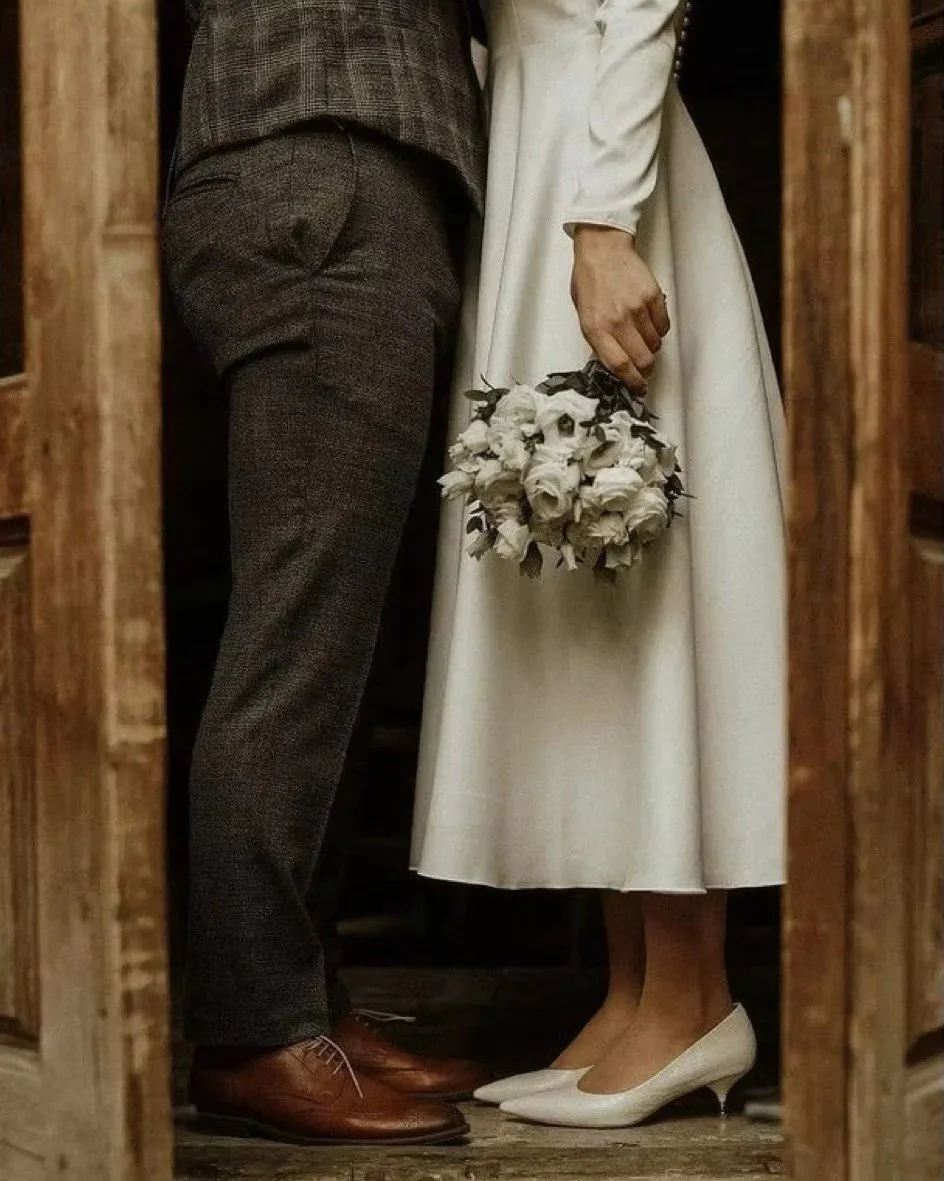 Close-up of a bride and groom standing close, holding a bouquet of white roses, with the bride in white heels and the groom in brown dress shoes, framed by wooden doorways, suggesting an intimate moment at a wedding.