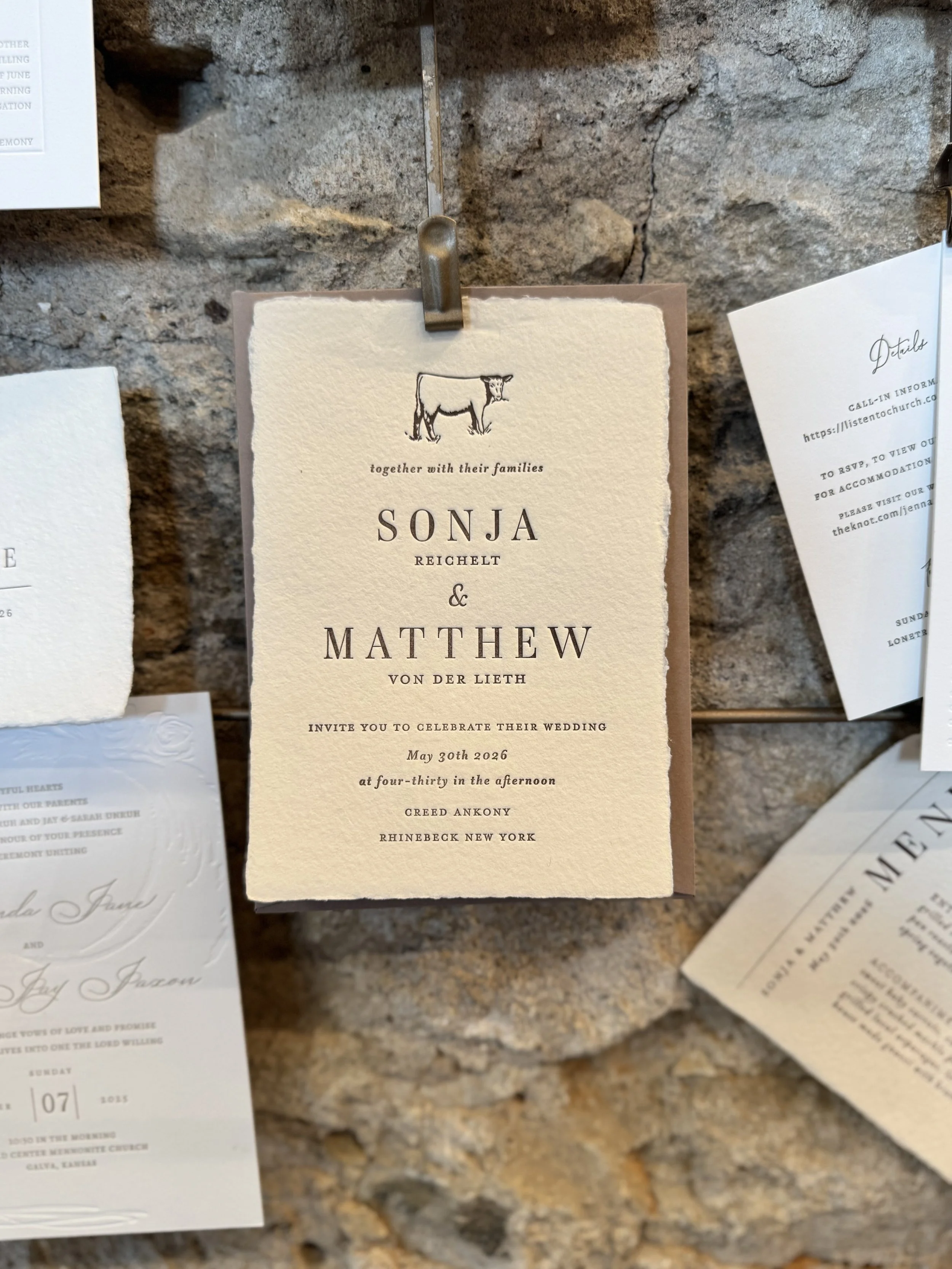 A wedding invitation card with a drawing of a cow, featuring the names Sonja Reichelt and Mathew, along with event details and date, displayed on a stone wall.