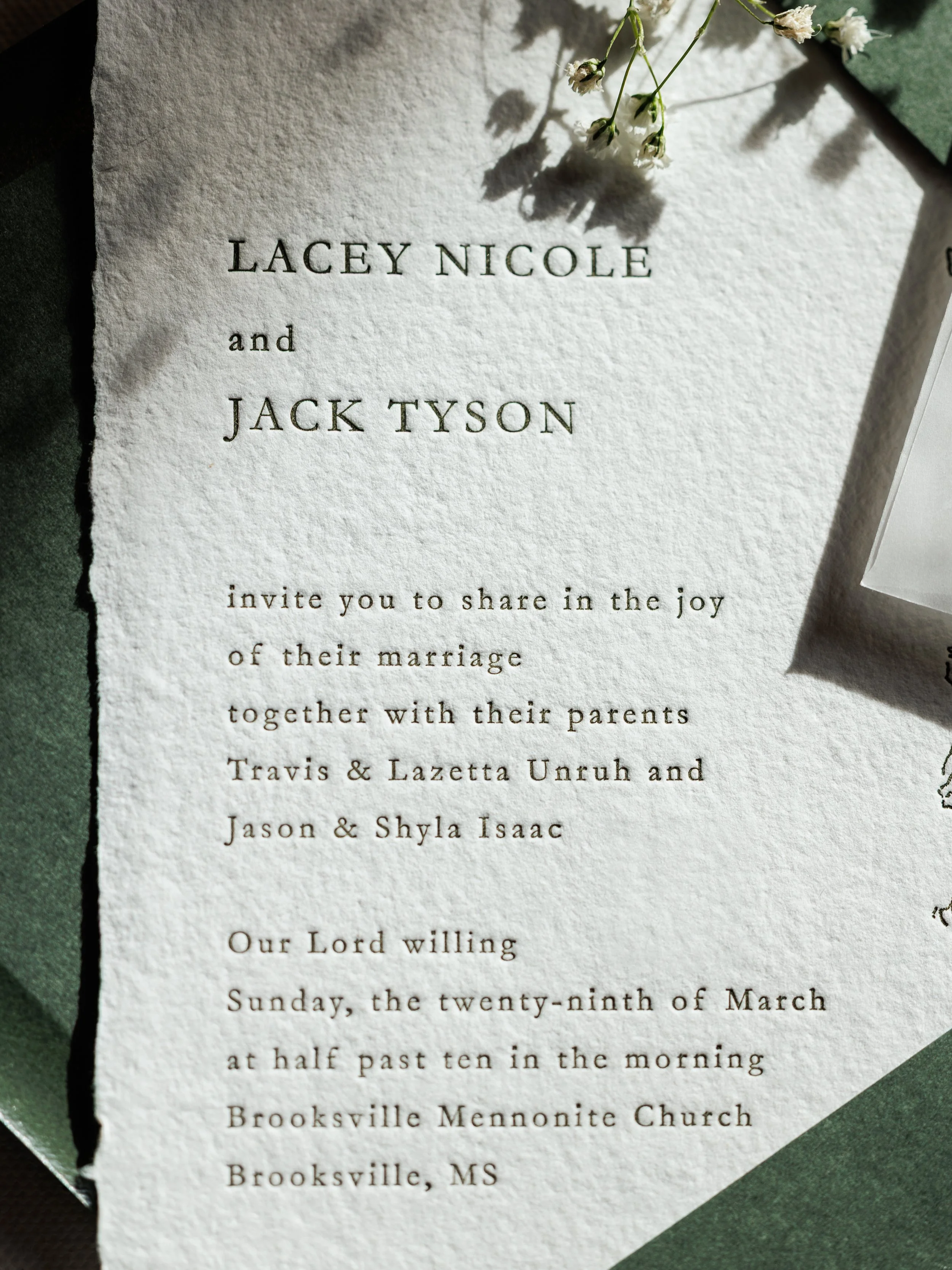 Close-up of a wedding invitation with elegant black text on textured white paper, decorated with small white flowers and paper leaves.