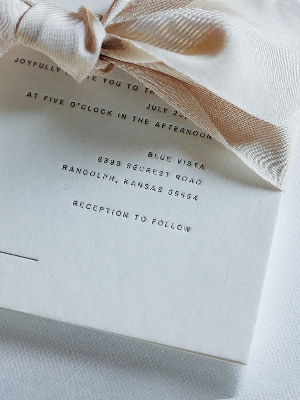 Close-up of a cream-colored invitation card with black text and a satin ribbon bow on top.