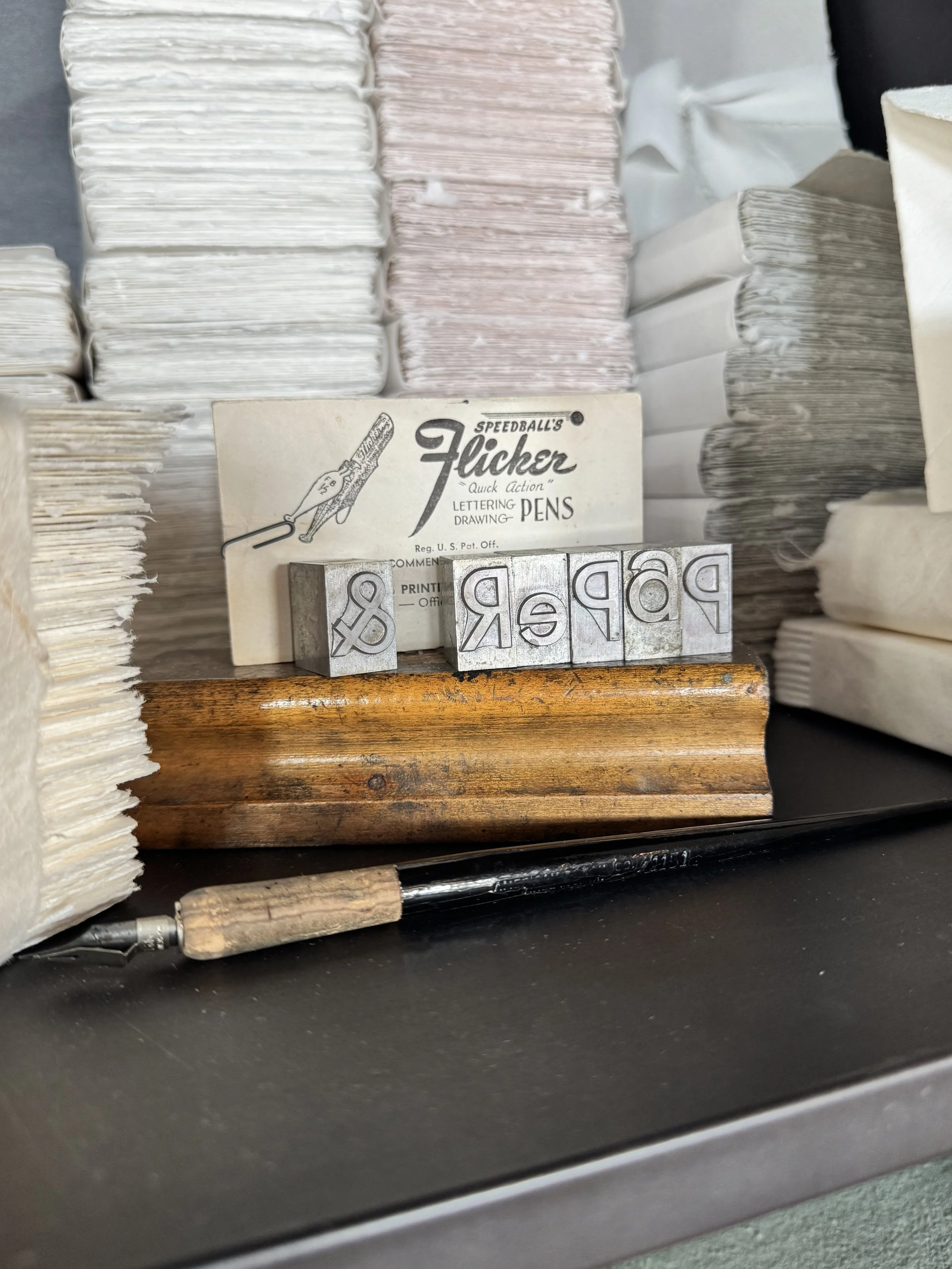 Stacks of old papers or envelopes, a vintage metal printing block spelling 'PRES' or similar, a Speedball Flicker lettering pen, and a black pen on a black surface.