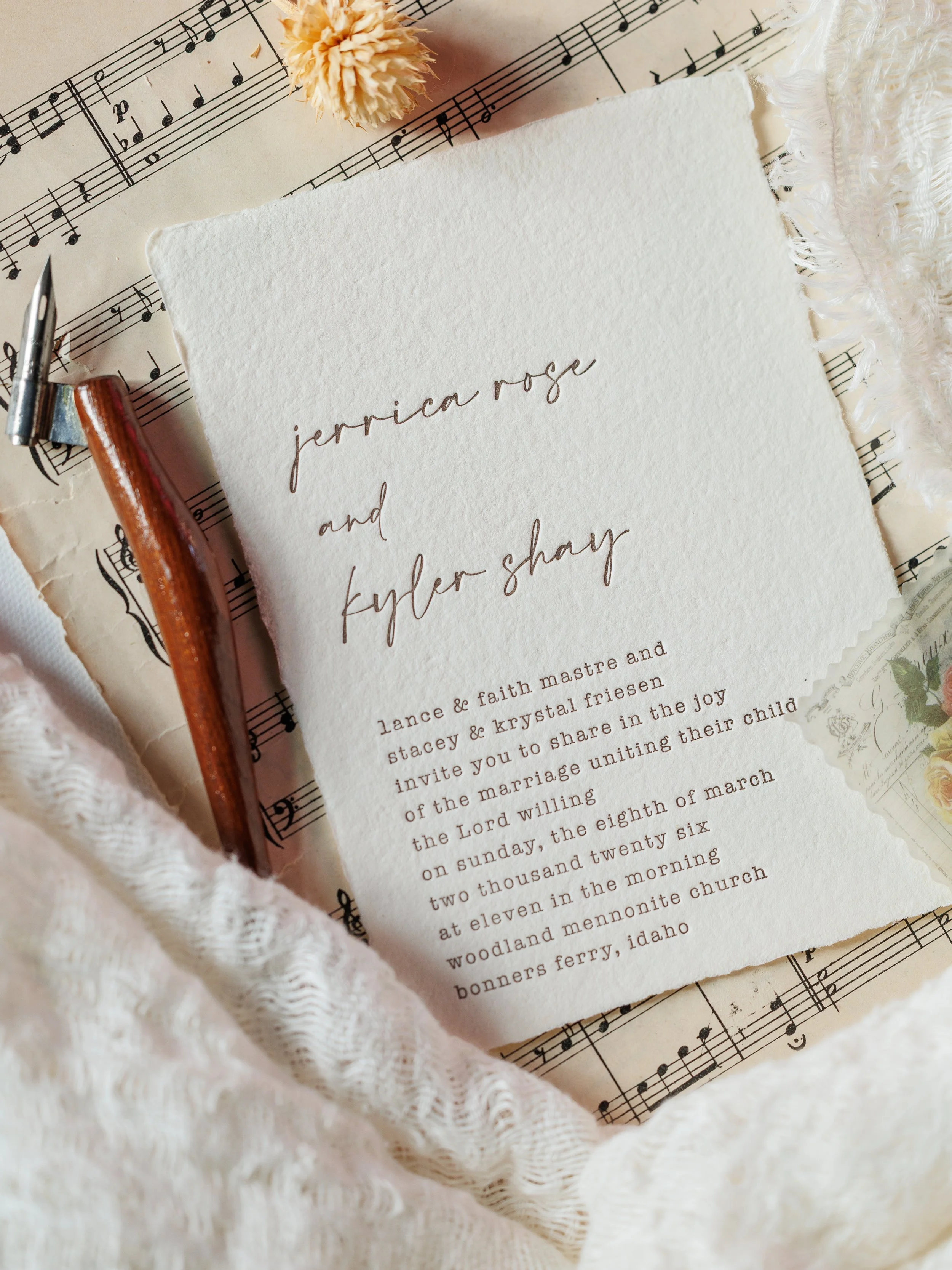 A wedding invitation with cursive writing, sitting on a sheet of music, with a soft pink pom-pom, a fountain pen, and a lace cloth nearby.