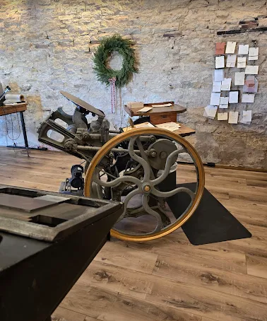 Vintage printing press with large wheel, set in a room with wooden floor, stone wall, and a holiday wreath in the background.