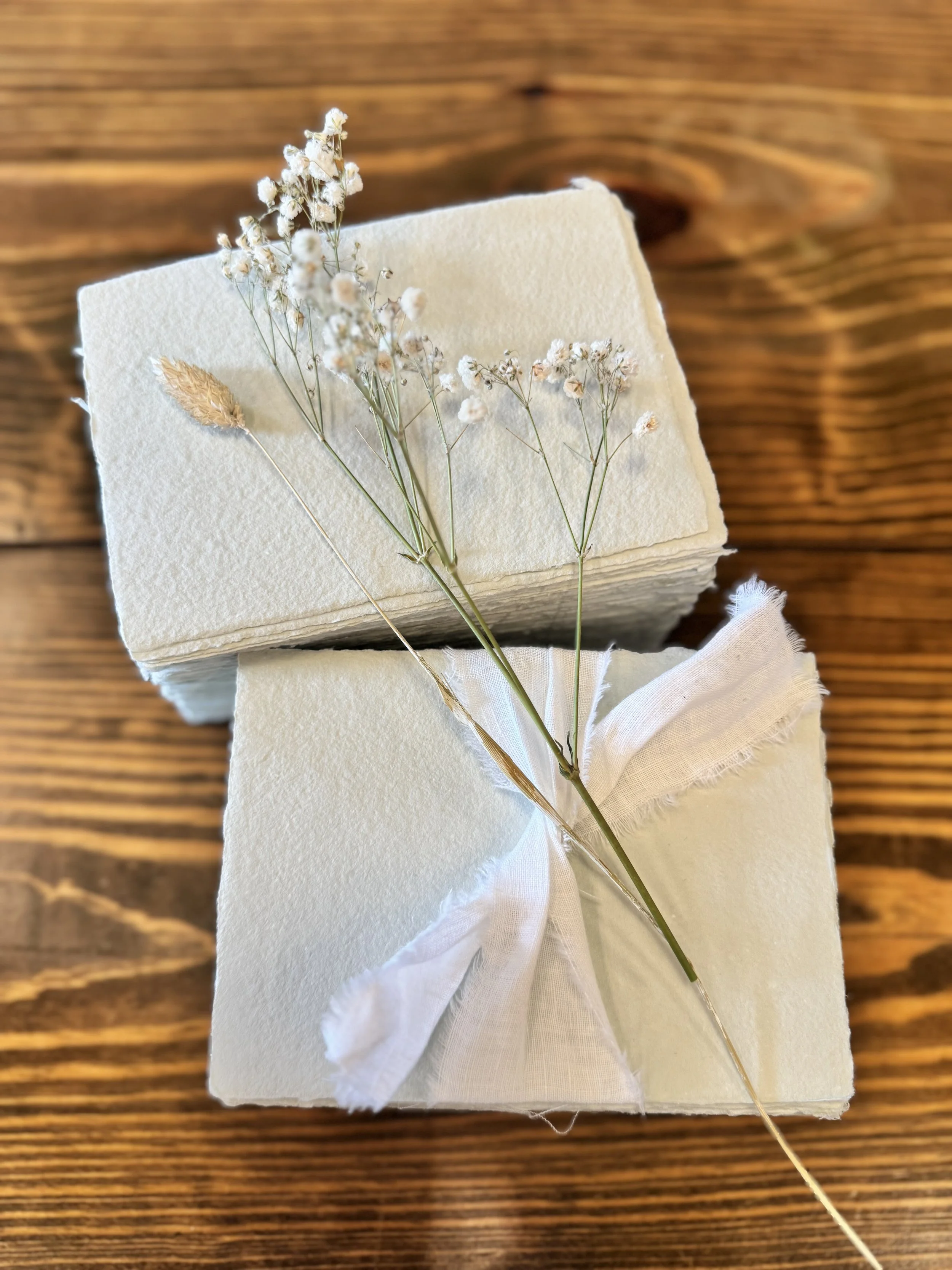 Small bundle of dried white baby's breath flowers attached to a white fabric and secured with a white ribbon, placed on a white textured surface, with a wooden background.