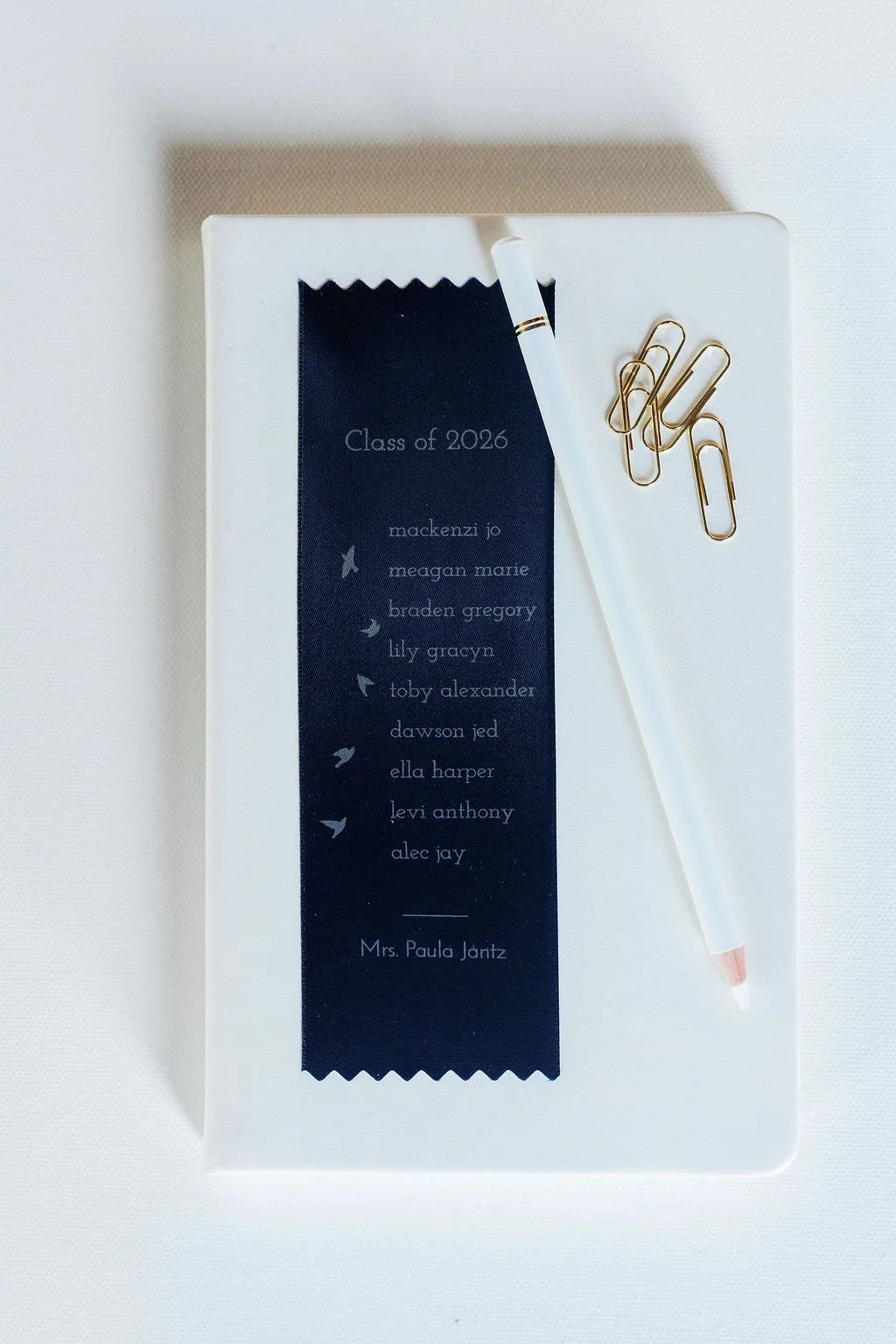 A white table with a white pen, gold paper clips, and a navy blue ribbon listing names of the graduating class of 2026, including Mrs. Paula Jantz.