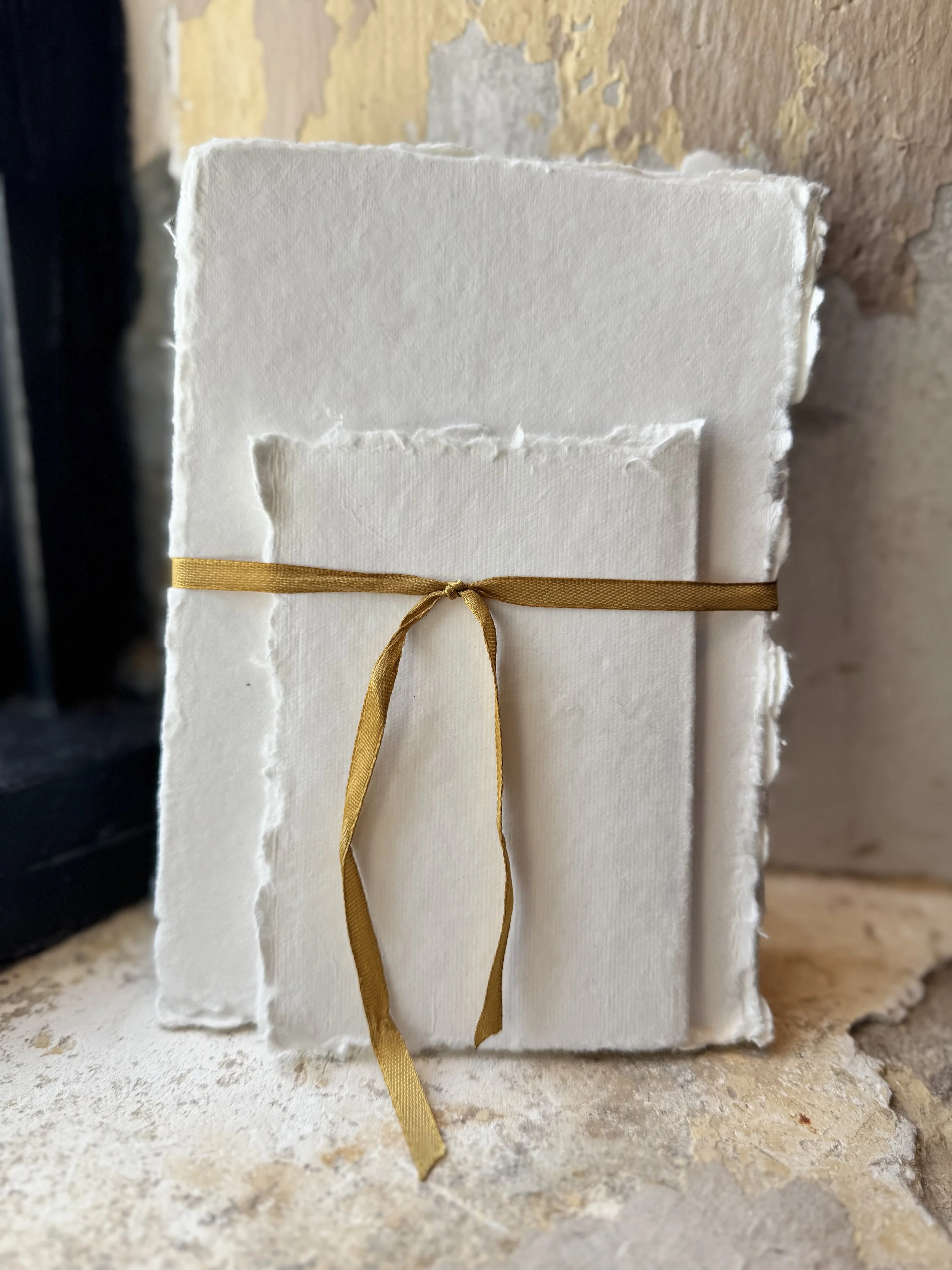 A close-up of a textured white paper with torn edges, wrapped with a gold ribbon tied in a bow, set against a rough, rustic wall.