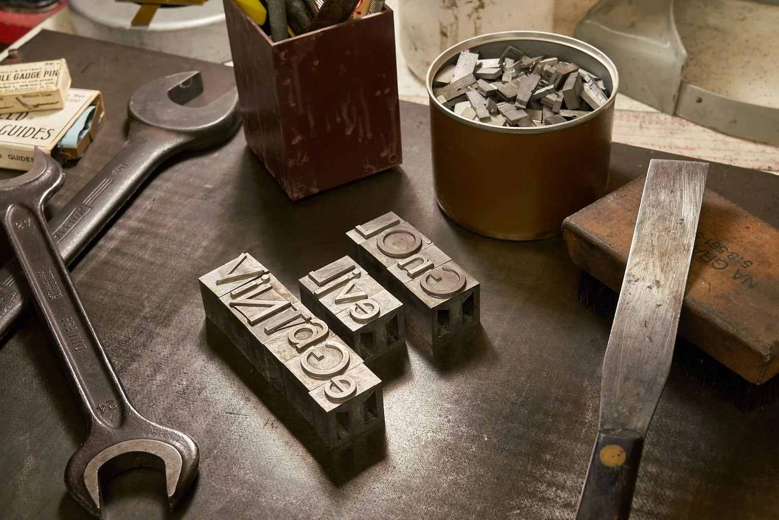 A workbench with metal letter stamps spelling 'CAVE' and 'GRA' alongside tools including a wrench, a small filing tool, a box of metal pieces, and a container of metal scraps.