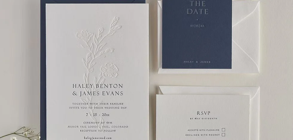 Wedding invitation suite including a main invitation card with embossed floral design and text, a navy blue RSVP card, and a white envelope, all arranged on a flat surface.