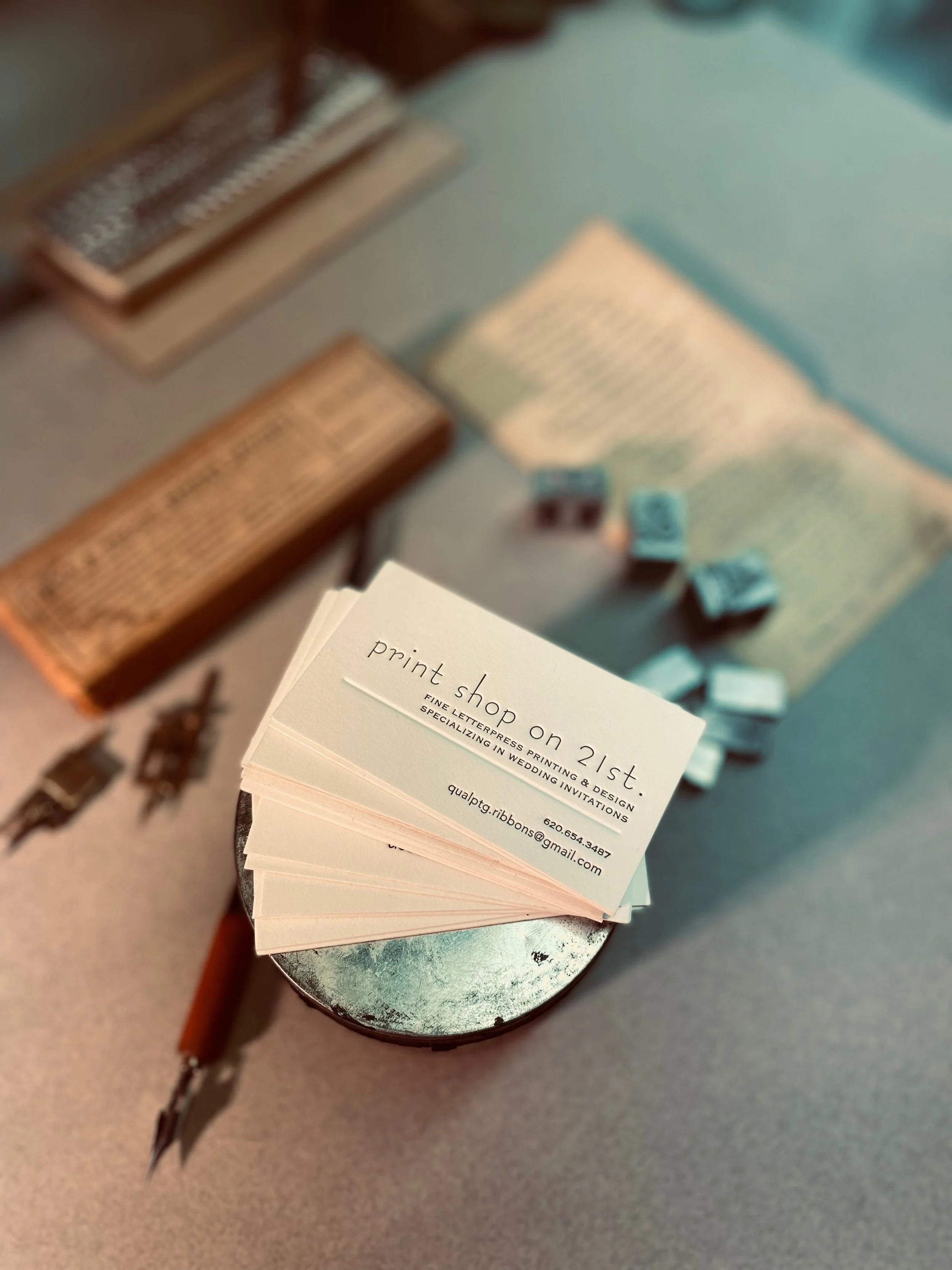 Business card for a printing company named 'Print Shop 21st' with contact information and services, placed on a table with blurred background items including small boxes and a piece of paper.