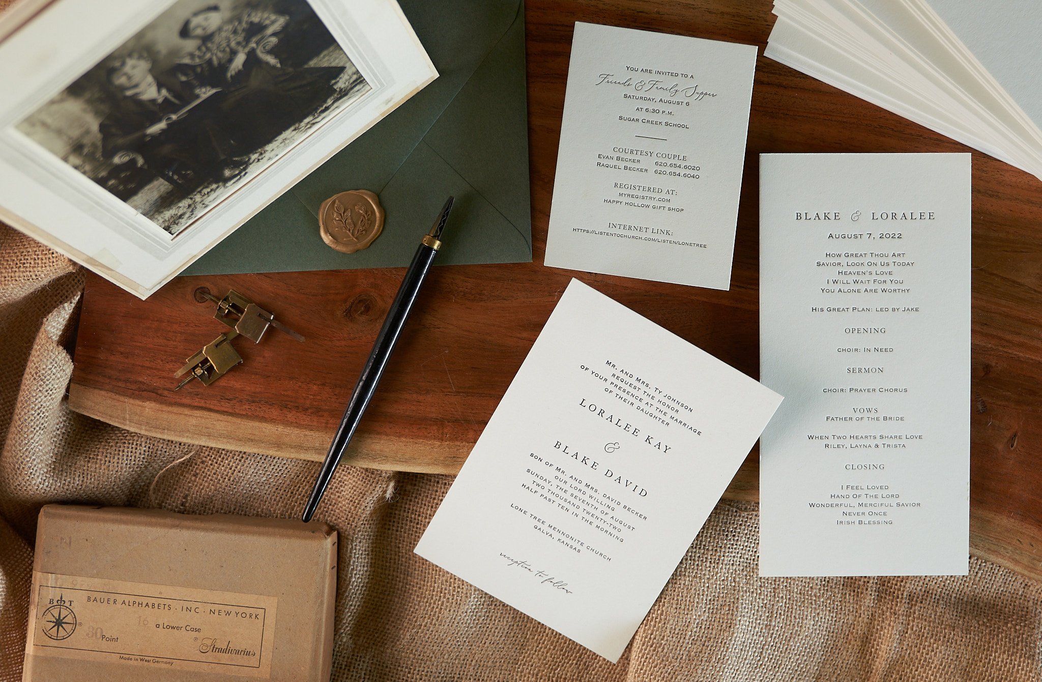 Wedding invitation cards, a sealed envelope with a wax stamp, a fountain pen, binder clips, a photograph, and an alphabet chart on a wooden surface and burlap fabric.