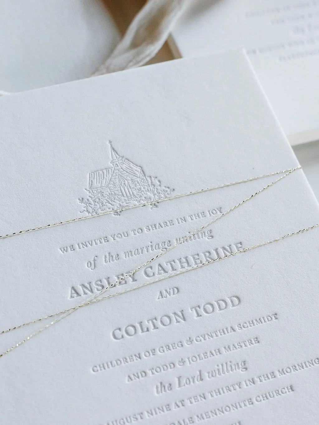 Close-up of a white wedding invitation with embossed and gold-embellished details, including an illustration of a church and text inviting guests to share in the wedding celebration of Ashley Catherine and Colton Todd.