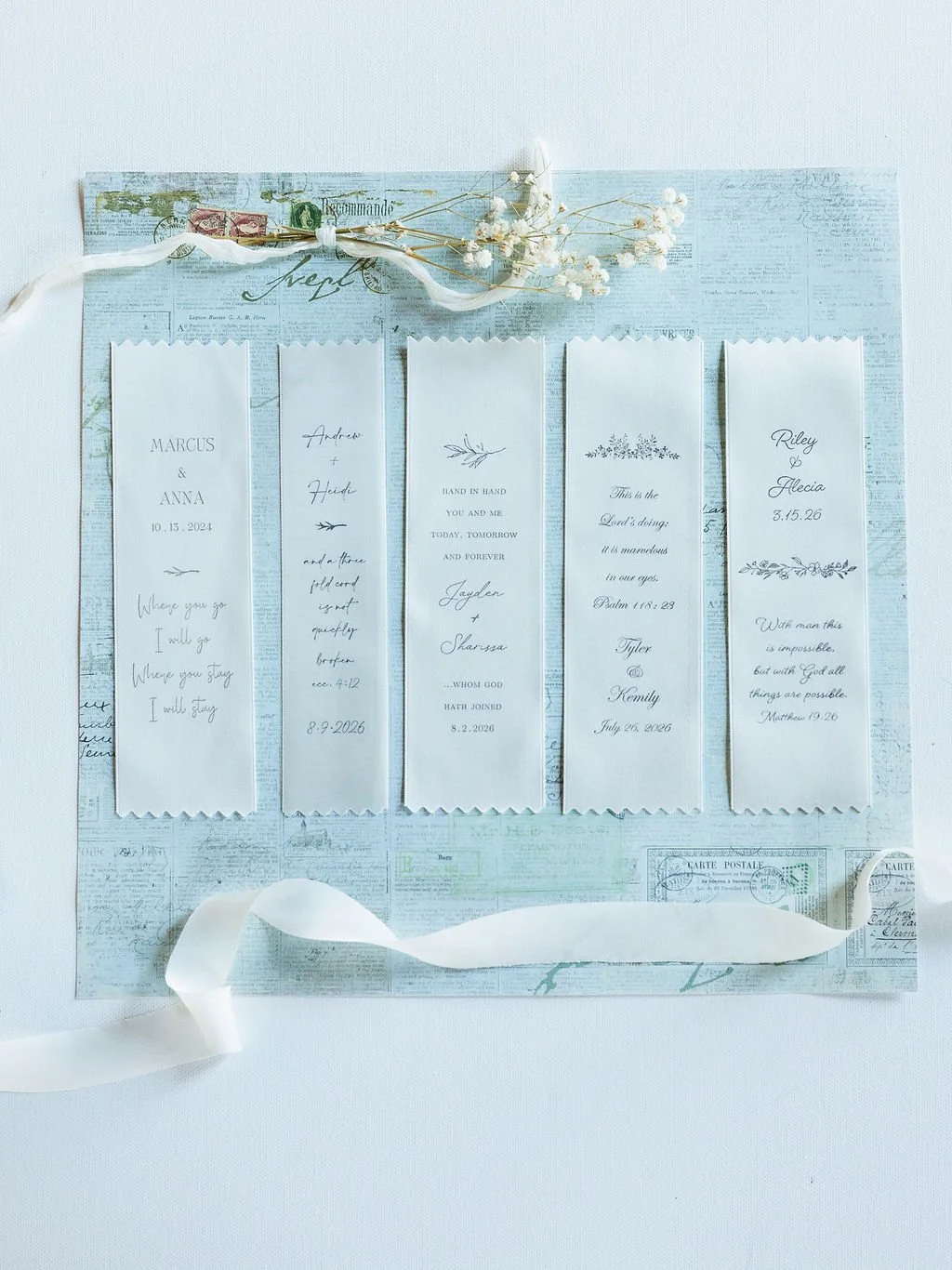 A white background with five celebrate bookmarks, each with handwritten names, dates, and quotes, decorated with white ribbon and baby's breath flowers, arranged on a vintage-style map paper.