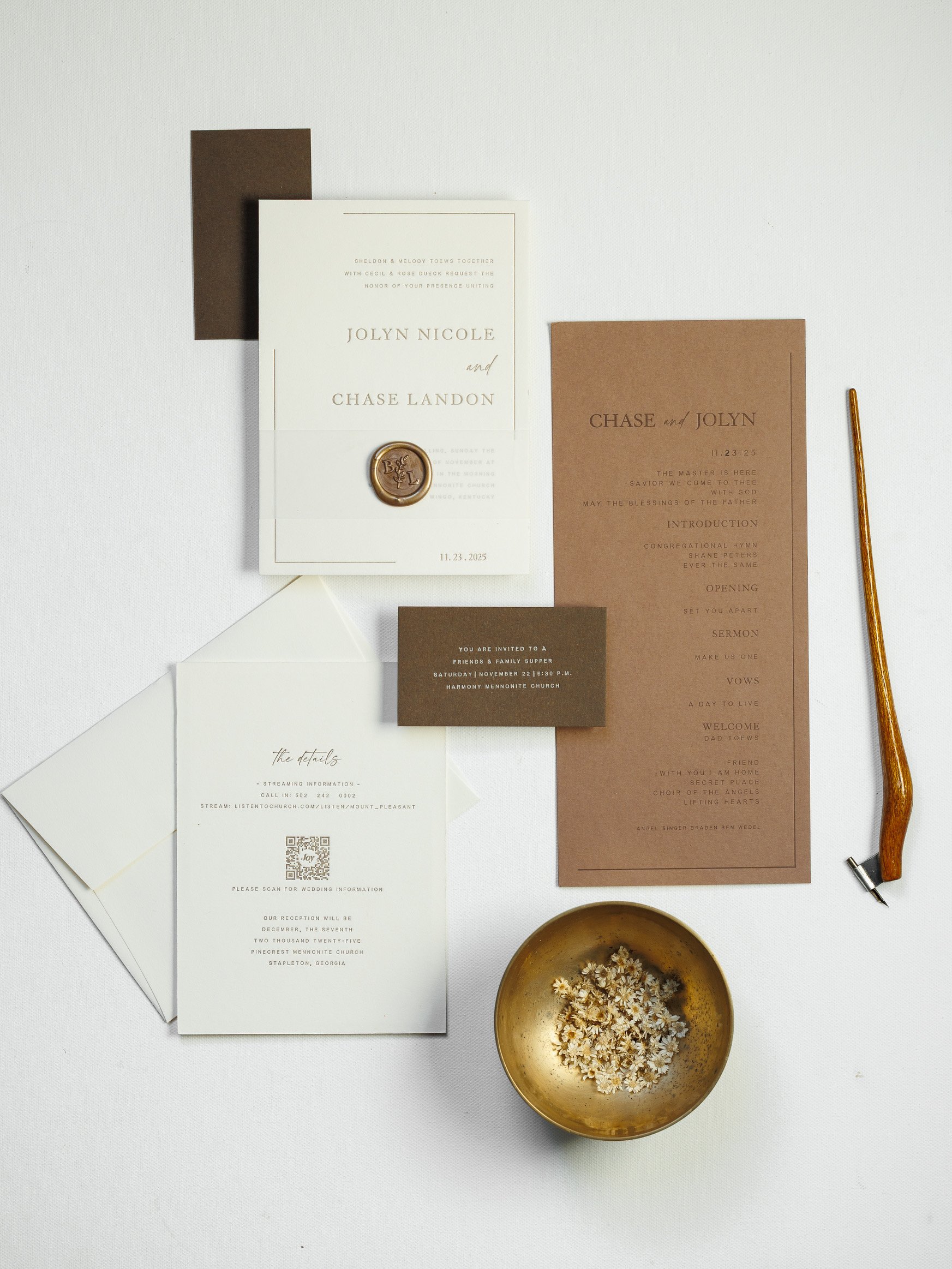 Wedding invitation suite with white and brown cards, a gold-emblem wax seal, a gold pen, and a bowl of small white flowers on a white surface.