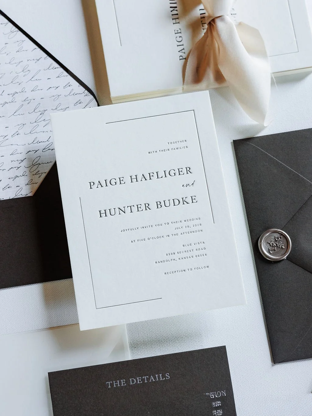 Wedding invitation card for Paige Haflinger and Hunter Budke with details of the ceremony on July 25, 2026, at Blue Vista, Randolph, Kansas, in black and white, accompanied by a black envelope with a wax seal and other stationery items.