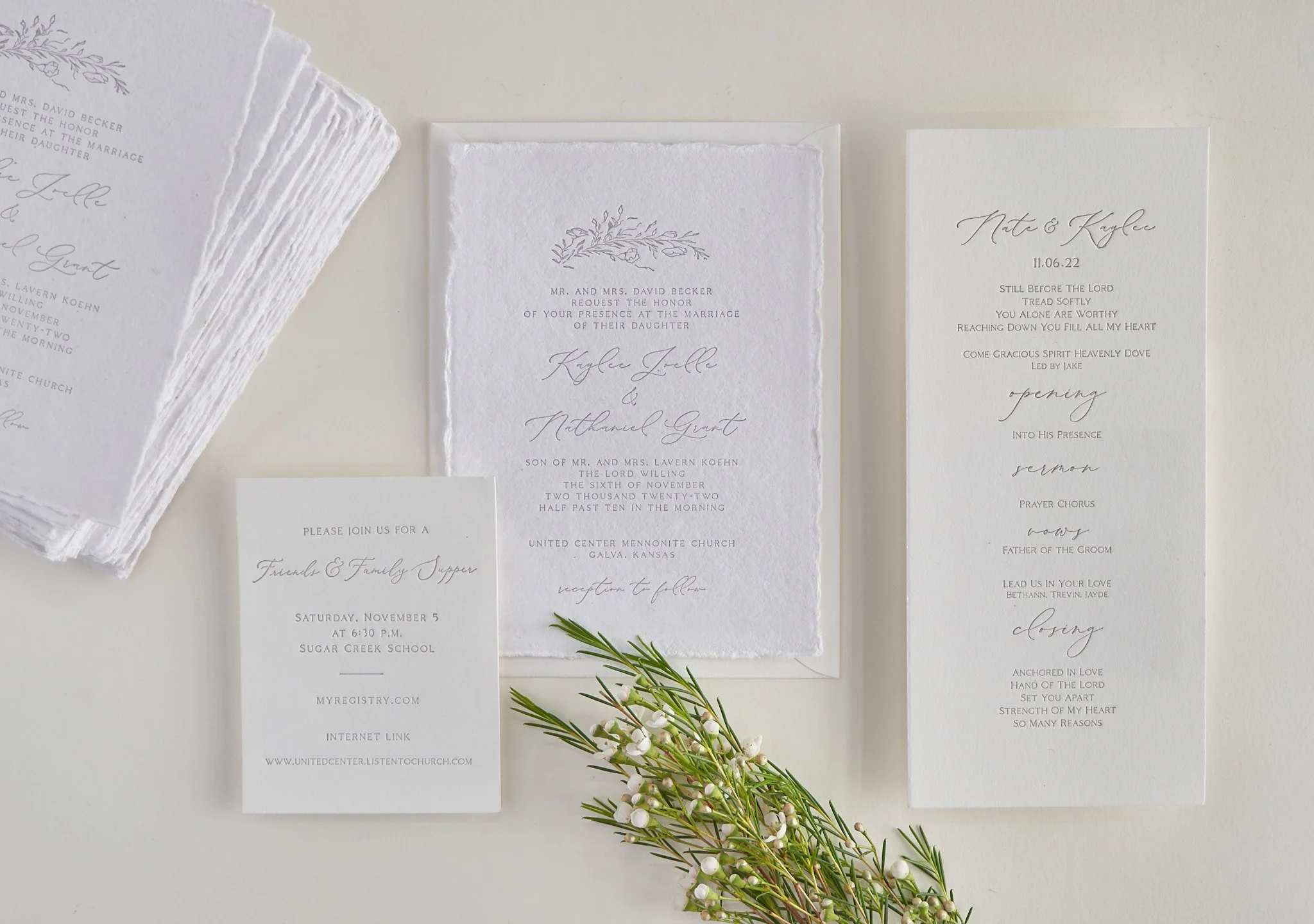 Elegant white wedding invitations with delicate floral and calligraphy design arranged on a white surface, complemented by a sprig of greenery and small white flowers.