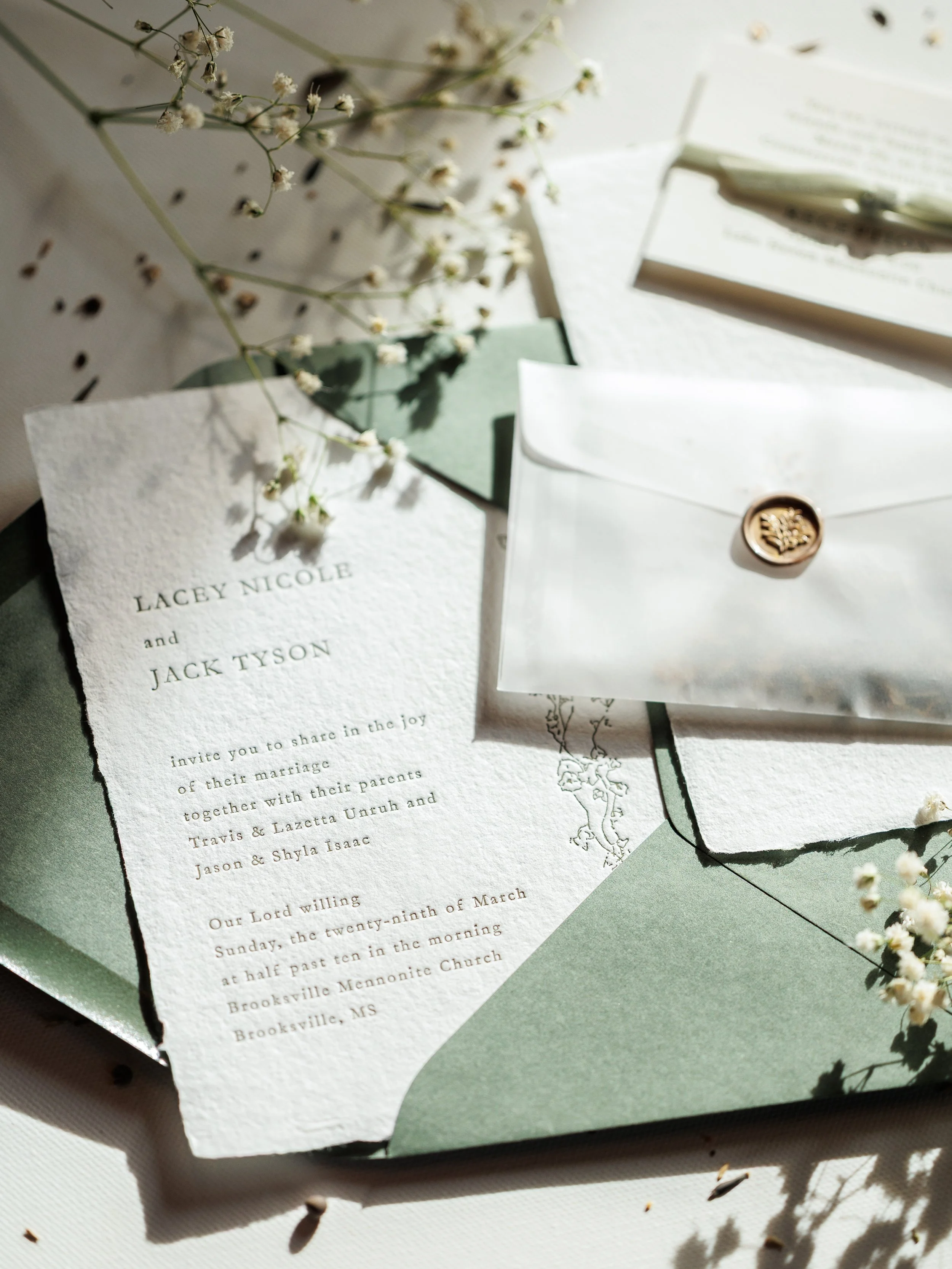 A wedding invitation card with text, a white envelope with a wax seal, sprigs of small white flowers, and scattered dark seeds or small objects on a light surface.