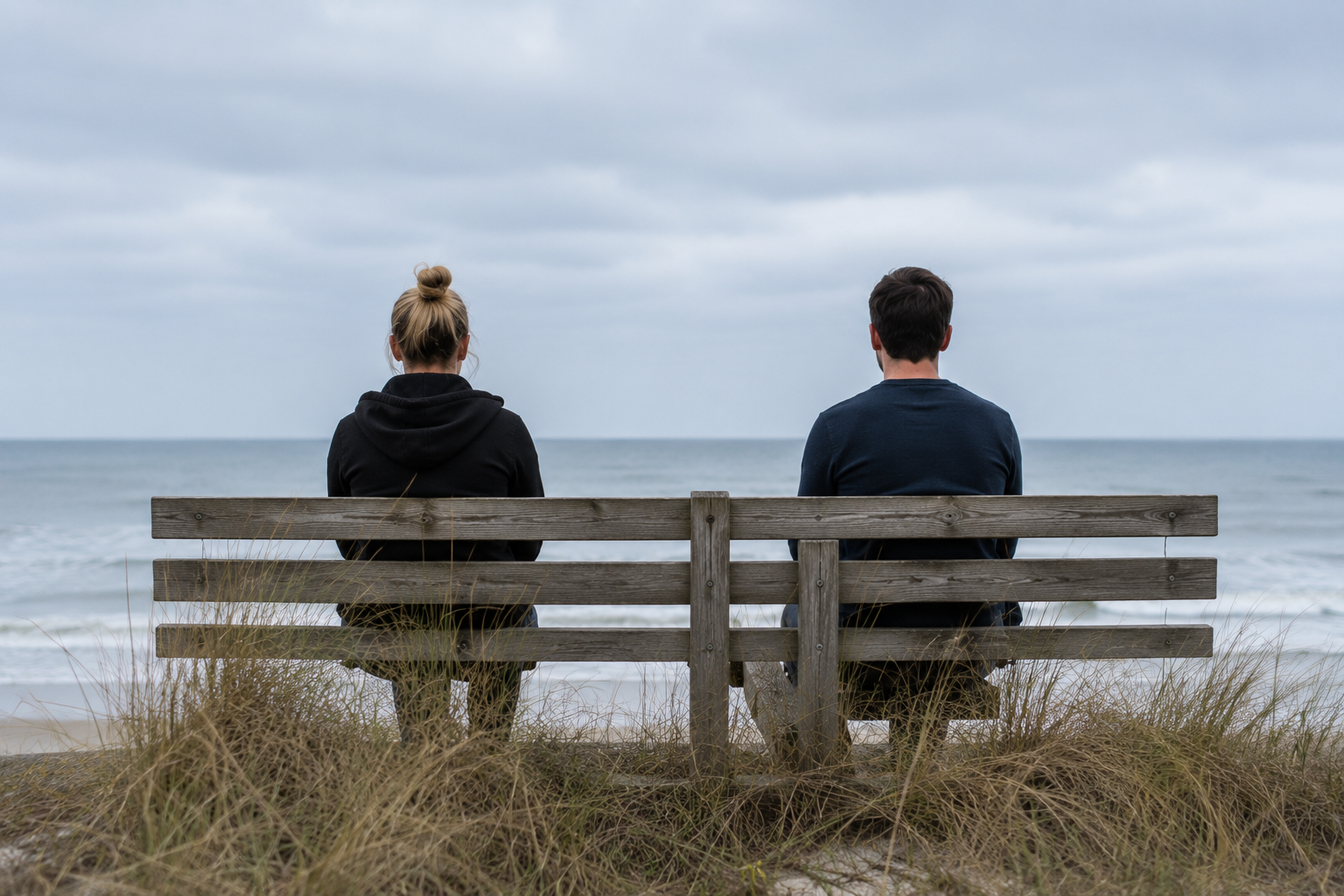 Two adults sitting apart on a bench overlooking the ocean, representing relationship challenges, emotional distance, and attachment patterns addressed in therapy.
