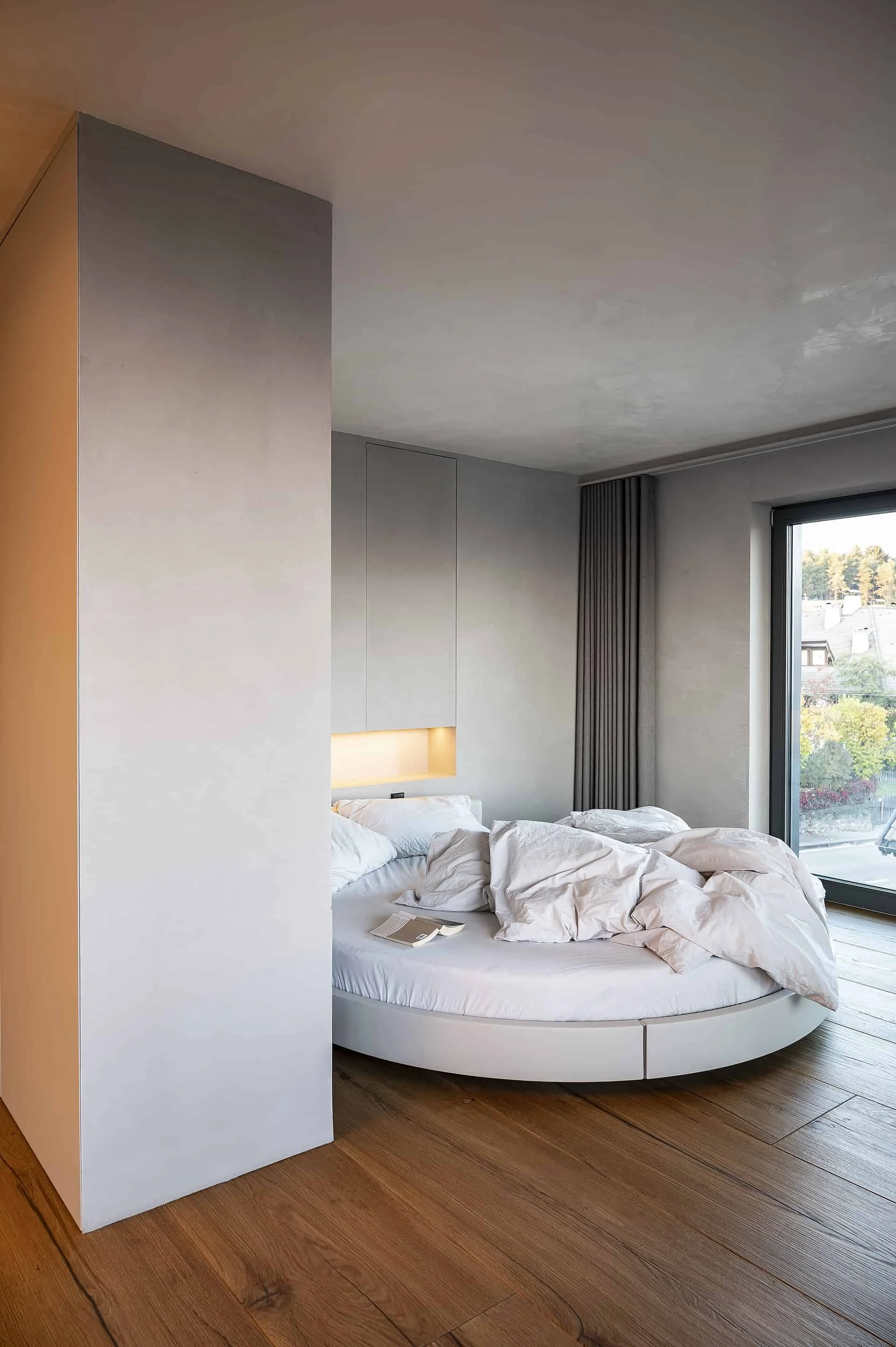 Ein modern eingerichtetes Schlafzimmer mit einem runden Bett, das unordentlich mit weißen Bettlaken und Kissen bedeckt ist. Es gibt eine große Fensterfront mit dunklen Vorhängen, durch die Tageslicht in den Raum fällt. Im Hintergrund sind eingebaute 