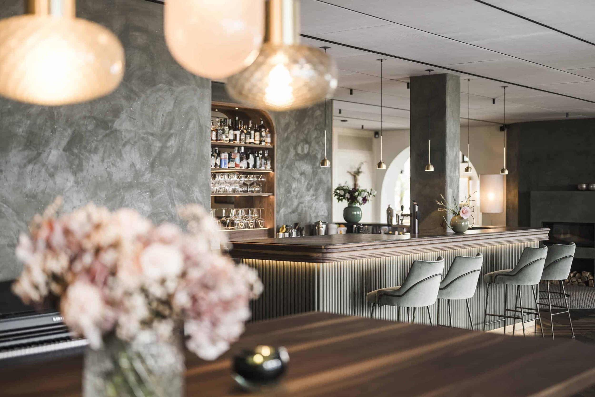 Ein modern gestalteter Bar- und Lounge-Bereich mit einer großen Bar, stilvollen hohen Stühlen, einer dekorativen Blumenvase auf dem Tisch, barista-ähnlichen Regalen mit Flaschen im Hintergrund und warmem, einladendem Licht.