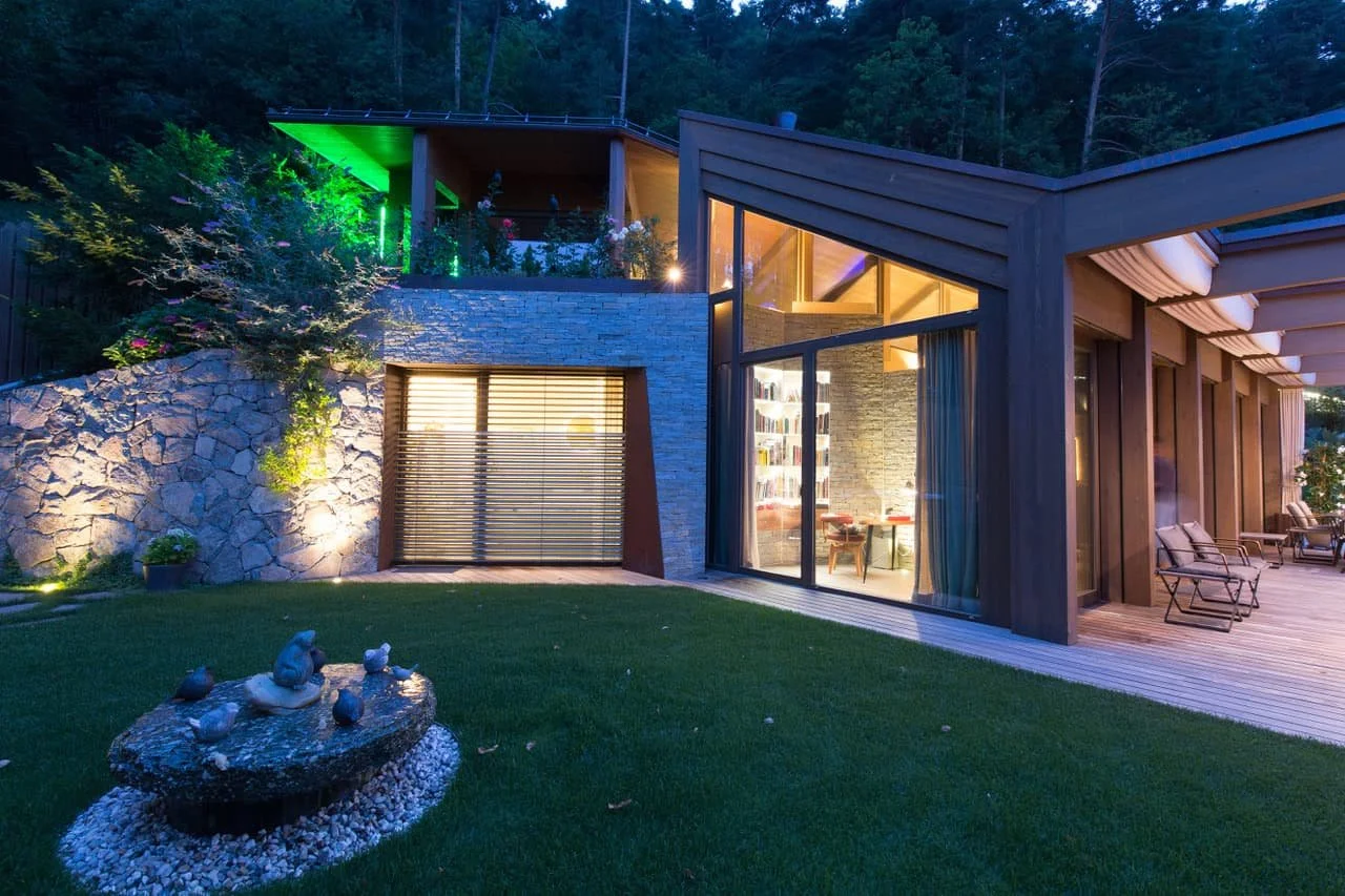 Modernes Haus mit großem Fenster und Terrasse, umgeben von einem Garten mit Steinfriedhof, auf einem Hügel mit Bäumen im Hintergrund bei Dämmerung, beleuchtet mit warmem Licht.