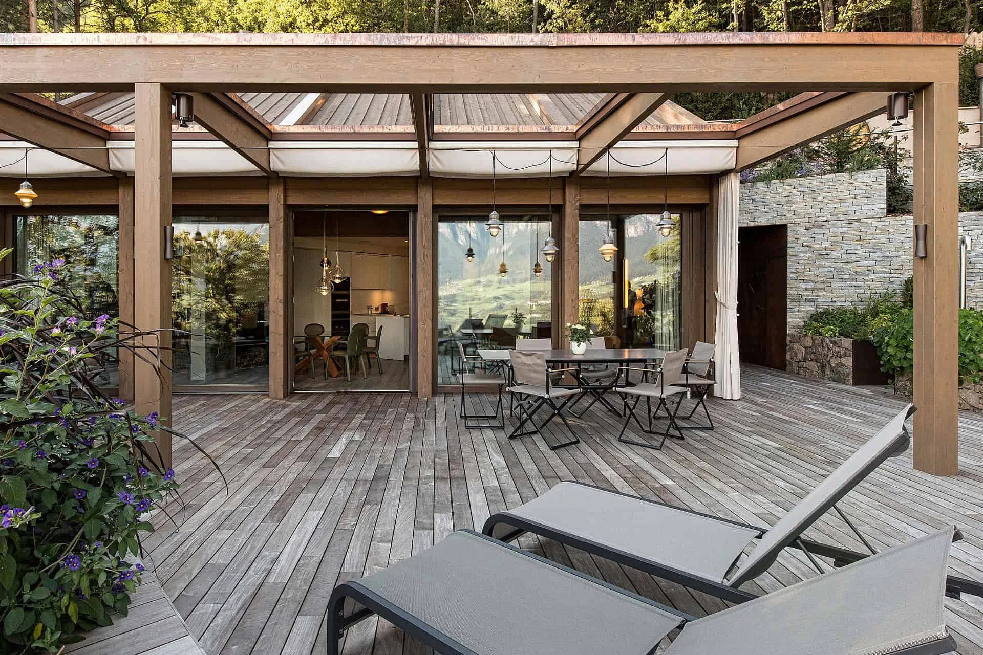 Holzdachterrasse mit Sitzmöbeln und Blick auf ein modernes Haus mit großen Glasfenstern, umgeben von Pflanzen und Bäumen.