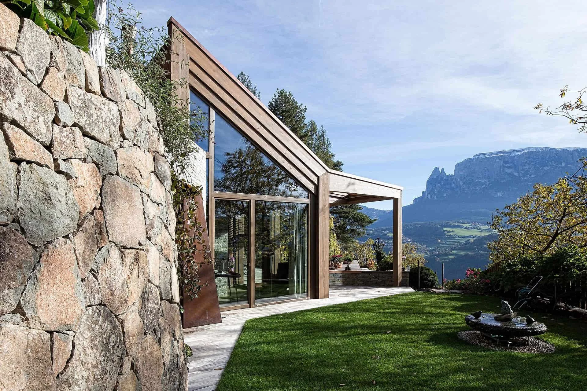 Modernes Haus mit großen Glasfenstern und Holzrahmen, umgeben von einem Garten mit grüner Wiese und Blick auf Berge im Hintergrund.