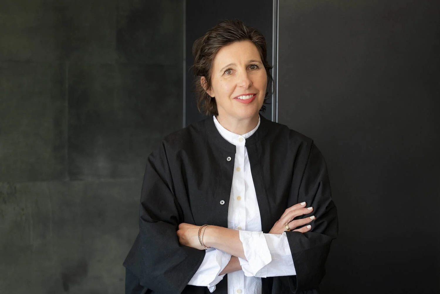 Architektin Angelika Mair steht vor einer dunklen Wand, lächelt leicht und hat die Arme verschränkt.
