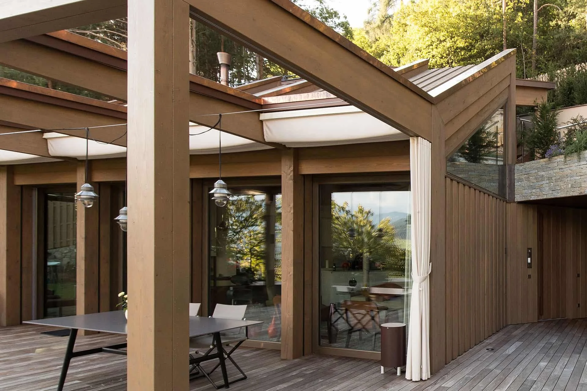 Moderne Terrasse mit Glaswänden, Holzdielenboden und viel Natur im Hintergrund. Es gibt einen Tisch mit Stühlen, eine weiße Vorhang und deko-Elemente.