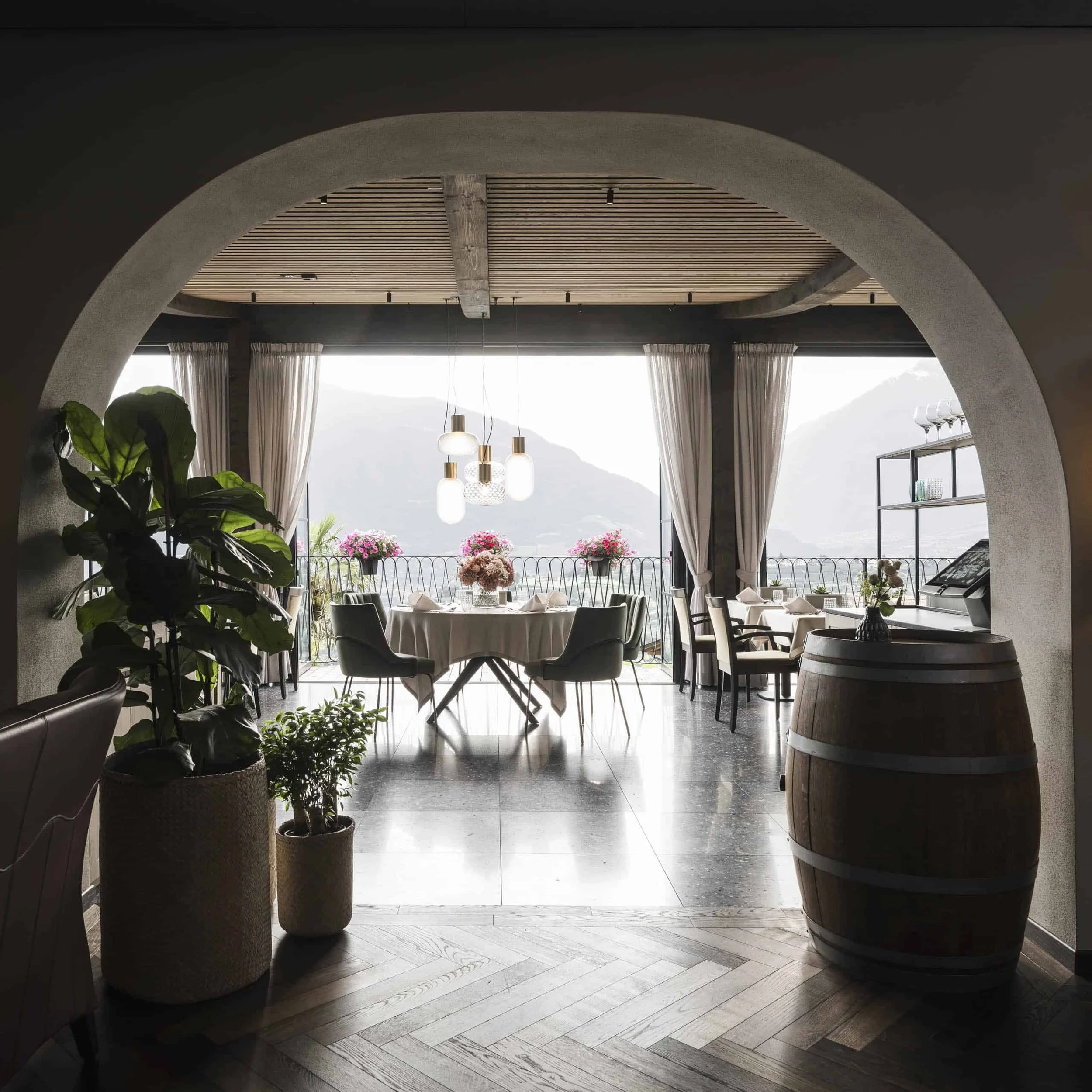 Blick in ein stilvolles Wohnzimmer mit großer Fensterfront, Pflanzen, modernen Lampen, Esstisch mit Blumen und pastellfarbenen Stühlen, Balkon mit Blumen, bergigem Ausblick im Hintergrund.