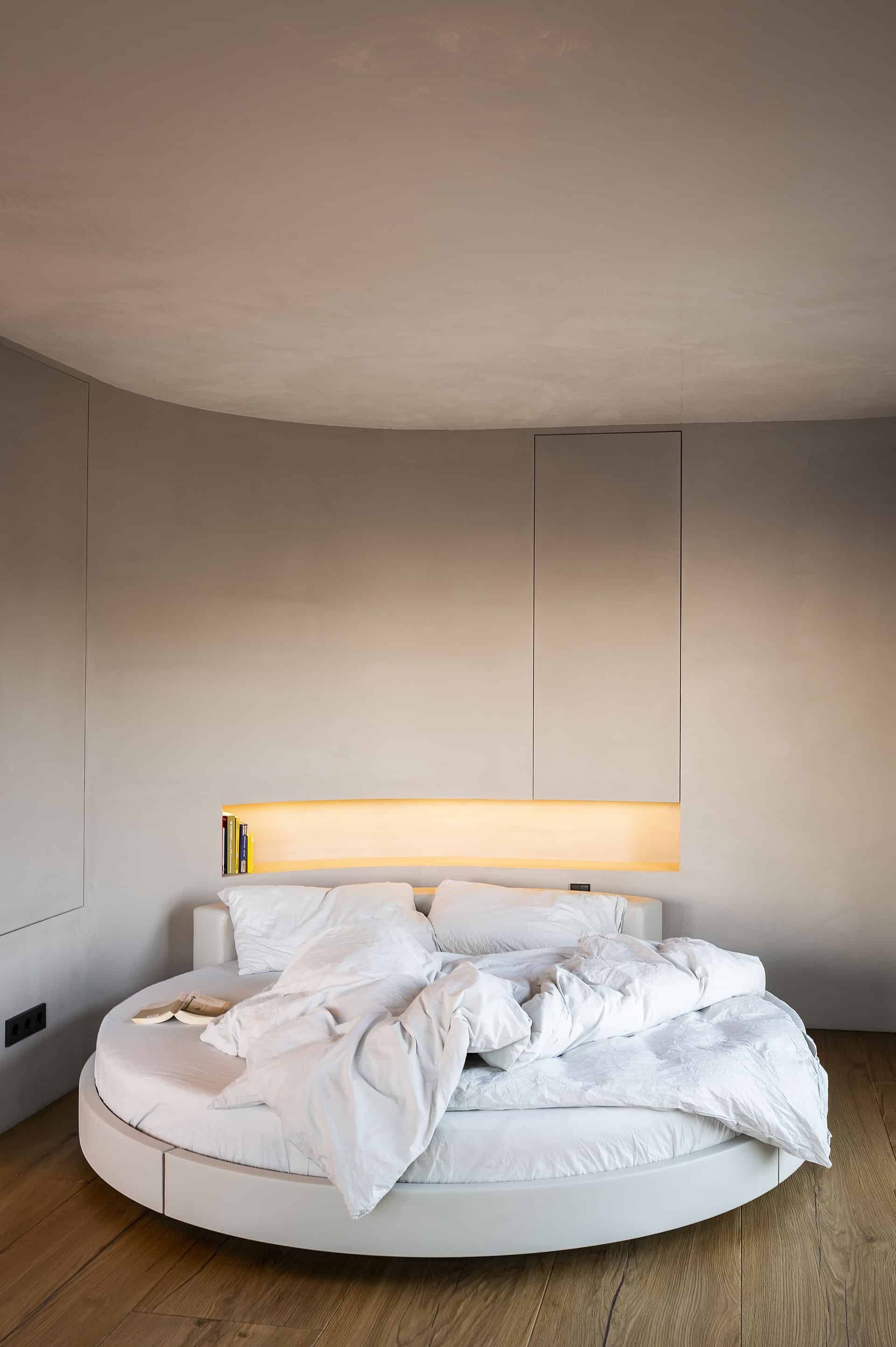 Ein unordentliches Bett mit weißen Laken und Kissen in einem modernen Schlafzimmer mit grauen Wänden und einer lampenartig beleuchteten Nische hinter dem Bett. Neben dem Bett liegt ein Buch.