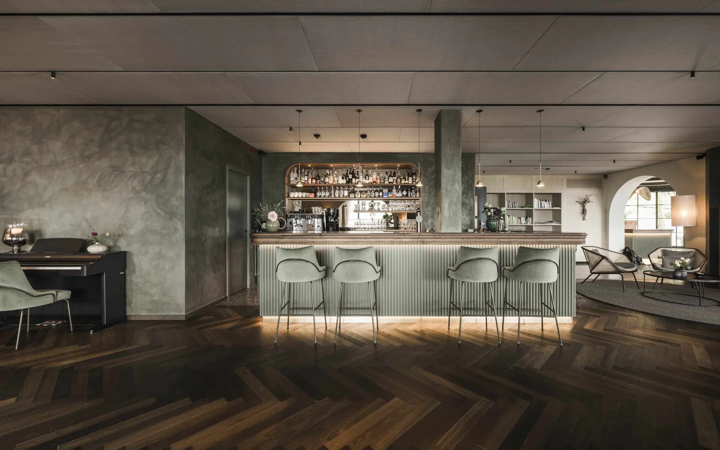 Ein modernes Bar- oder Lounge Bereich mit Theke, Barhockern, Vitrinen mit Spirituosen, einem Piano, einem gemütlichen Sitzbereich mit Sesseln und Leseregalen, viel natürlichem Licht durch große Fenster.