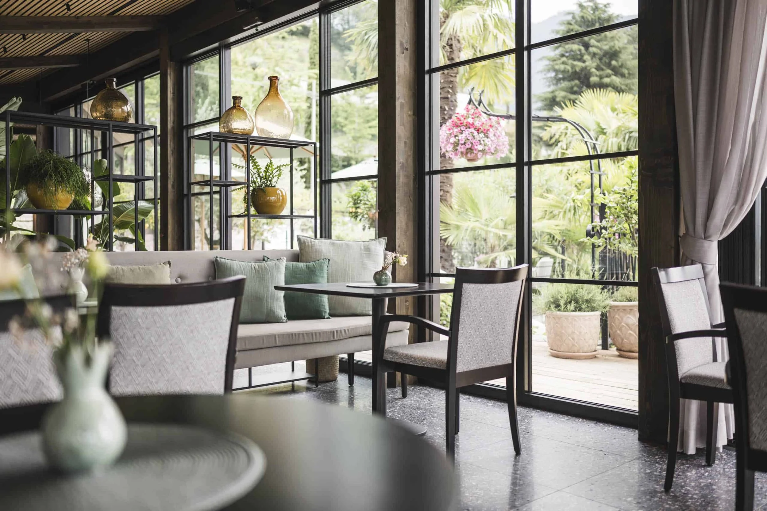 Innenraum eines modernen Restaurants mit großen Fenstern, bequemen Stühlen und Dekorationsgegenständen wie Vasen und Pflanzen. Draußen sind Palmen und bunte Blumen zu sehen.