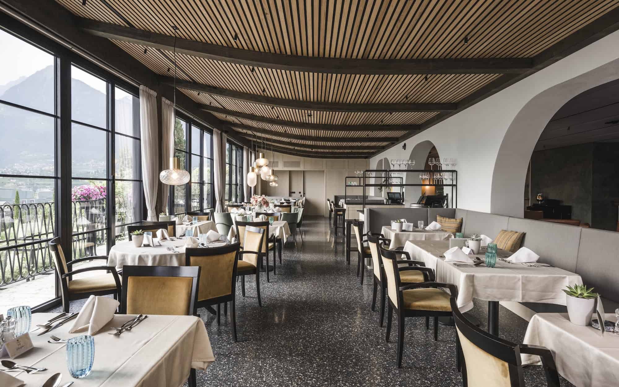 Innenansicht eines eleganten Restaurant mit großen Fenstern, weißen Tischtüchern, Stühlen und moderner Beleuchtung.