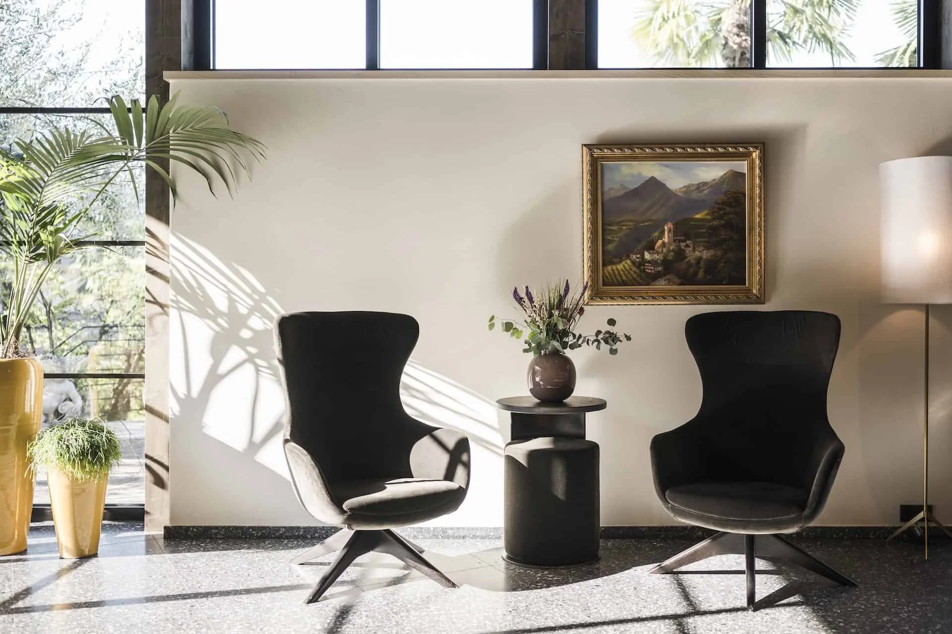 Ein stilvoller Sitzbereich mit zwei schwarzen Sesseln, einem runden Tisch mit einer Vase mit Lavendel und Eukalyptus, einem Gemälde eines Bergdorfes an der Wand, eine Stehlampe und grüne Pflanzen vor Fenstern mit Blick auf die Natur.