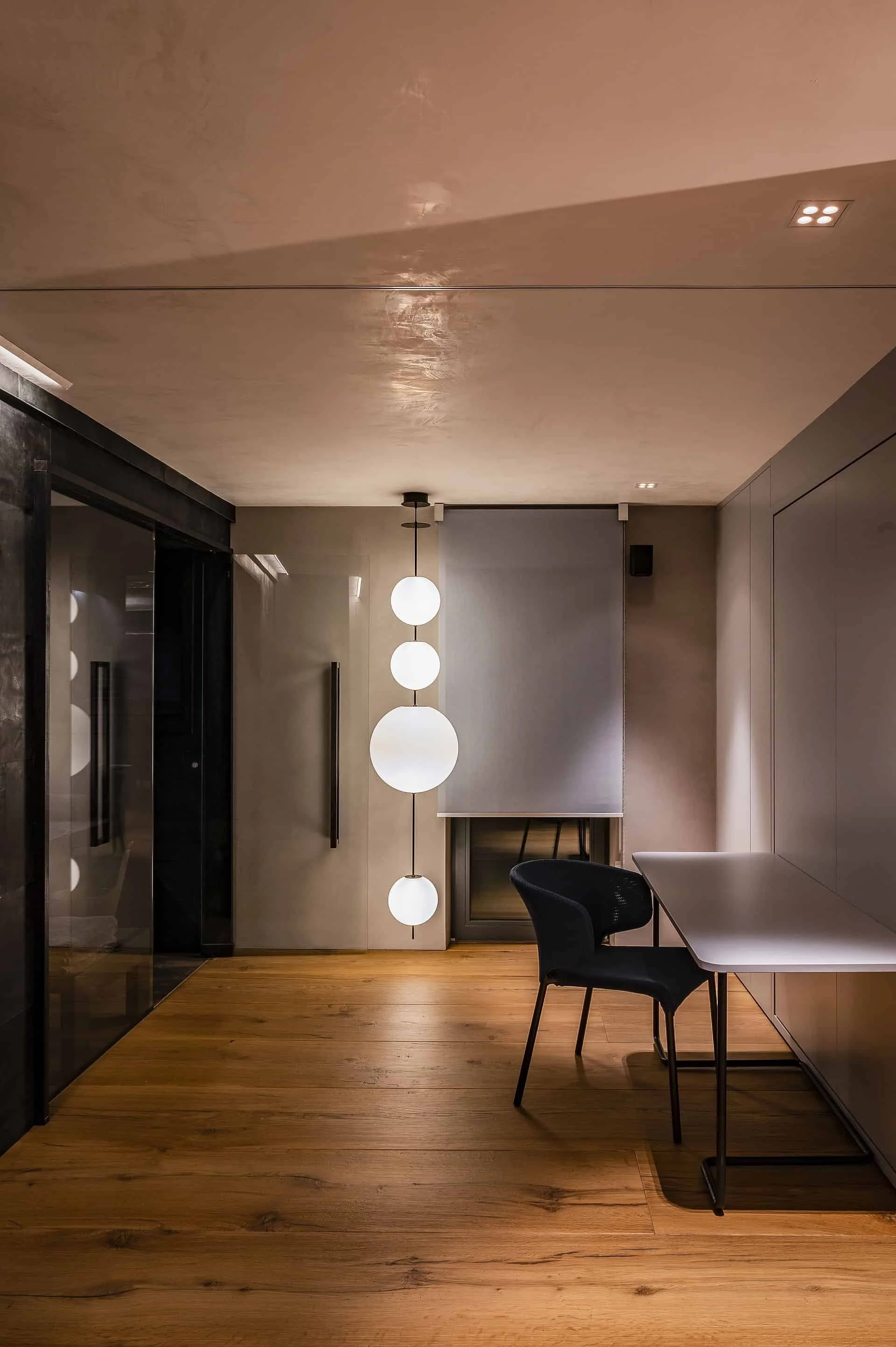 Innenaufnahme eines modernen Raums mit Holzboden, einem Tisch, einem schwarzen Stuhl, einer Lampenkette mit kugelförmigen Lampen und Fliegengittern an den Fenstern.