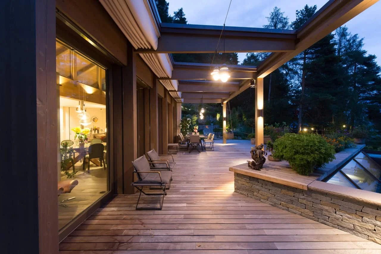 Ein abgeschiedener Balkon mit Holzdielen, umgeben von Bäumen, beleuchtet bei Dämmerung, mit Gartenmöbeln, Pflanzen und Dekorationen.