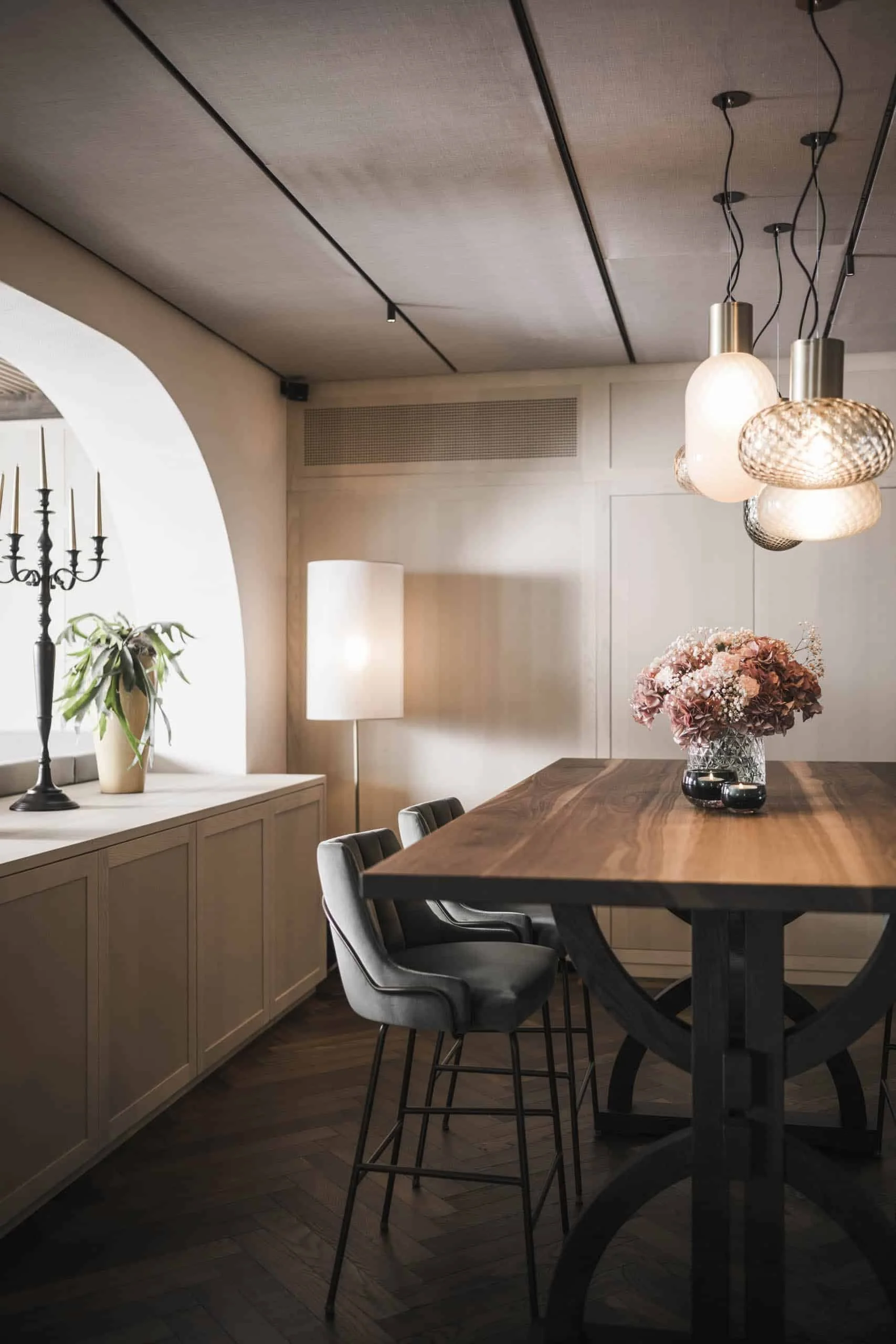 Innenansicht eines modernen Esszimmers mit hellem Holz, einem großen Holztisch, sechs grauen Stühlen, einer weiße Stehlampe, einem Blumenarrangement auf dem Tisch, einem weißen Vorhang, Wandpaneelen und einer Beleuchtung aus mehreren modernen Lampen