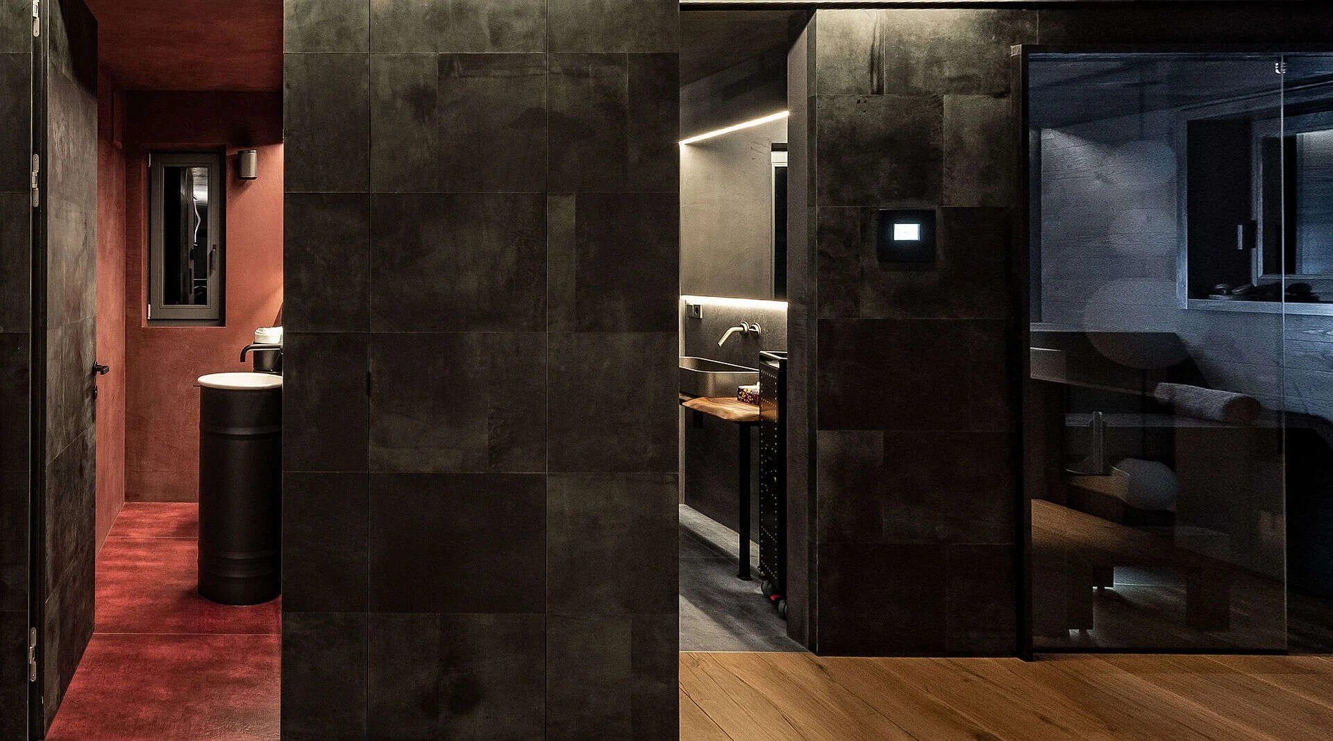 Innenansicht einer modern gestalteten Unterkunft, vermutlich eine Hotelzimmer- oder Apartment-Einrichtung, mit dunklen Wänden, einem kleinen Küchenbereich und einem Schlafzimmer im Hintergrund.