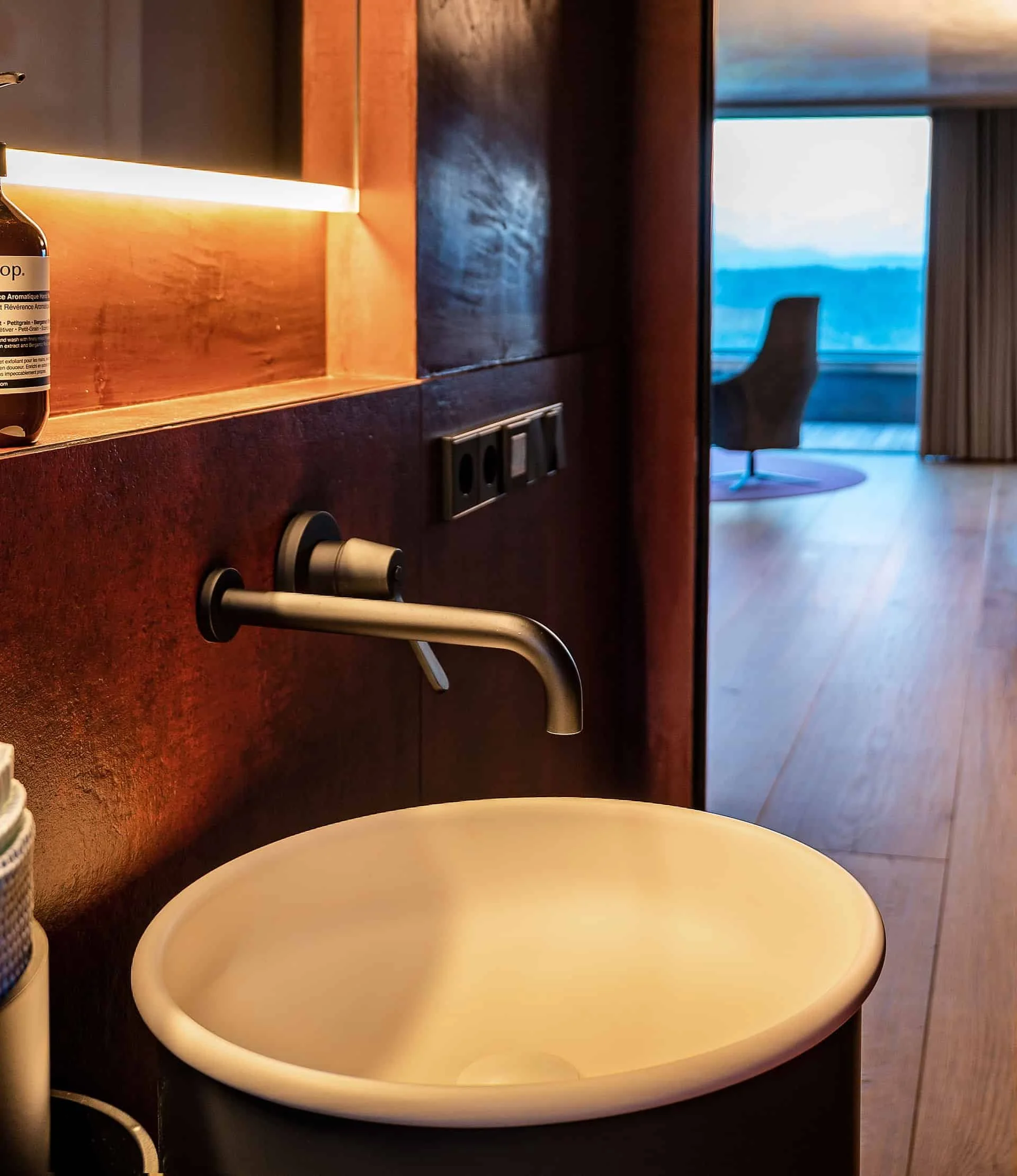 Ein runder, weißer Waschbecken mit schwarzem Hintergrund, eine Wasserhahn, eine Flasche auf der linken Seite und ein Stuhl im Hintergrund vor einem Fenster mit Blick auf eine Landschaft am Horizont.
