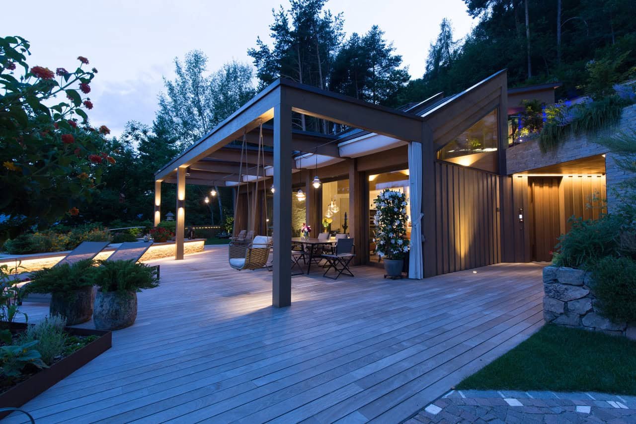 Ein moderner Balkon im Abendlicht mit Gartenmöbeln, Pflanzen und Holzdecke, umgeben von Bäumen.