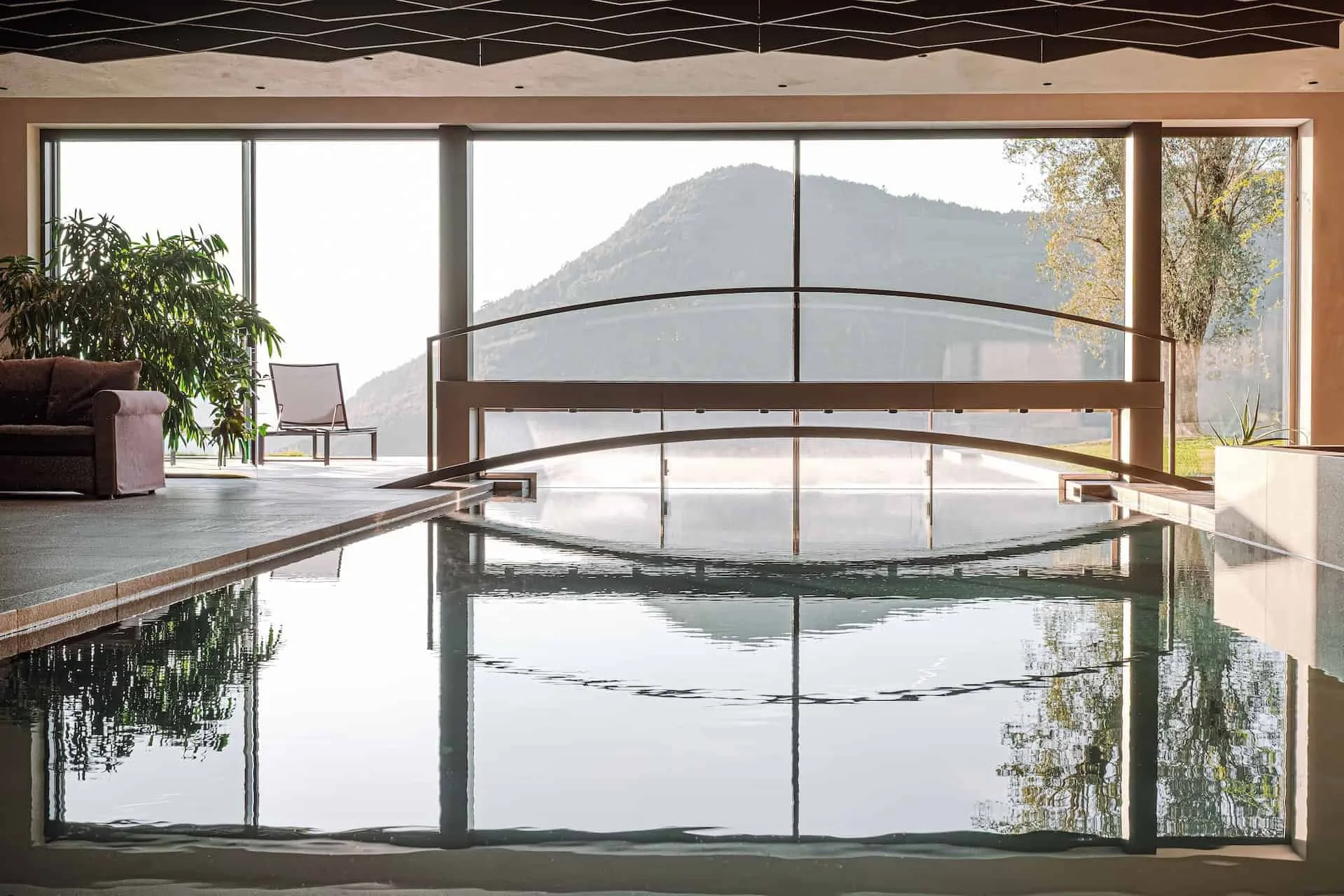 Innenansicht eines Spa- oder Thermalbads mit Blick auf eine bergige Landschaft durch große Fenster, Wasserbecken im Vordergrund, einige Pflanzen und Sitzgelegenheiten auf der linken Seite.