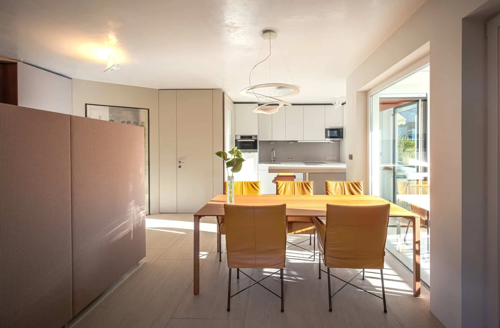 Offenes Wohnzimmer mit Moderner Einrichtung, hellem Holz Tisch, gelbe Stühle, große Fenster mit Balkon, Küche im Hintergrund, Pflanzen auf dem Tisch, Deckenleuchte. Nachhaltige, minimalistische Inneneinrichtung.