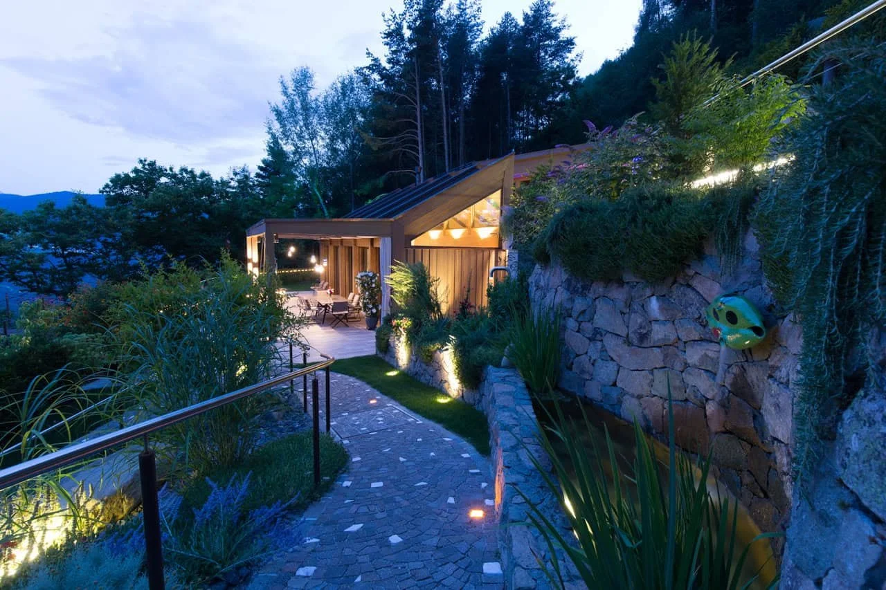 Abendansicht eines modernen Hauses mit beleuchtetem Garten, Steinpfad, viel Grün und einer Terrasse im Hintergrund, umgeben von Bäumen in bergiger Umgebung.