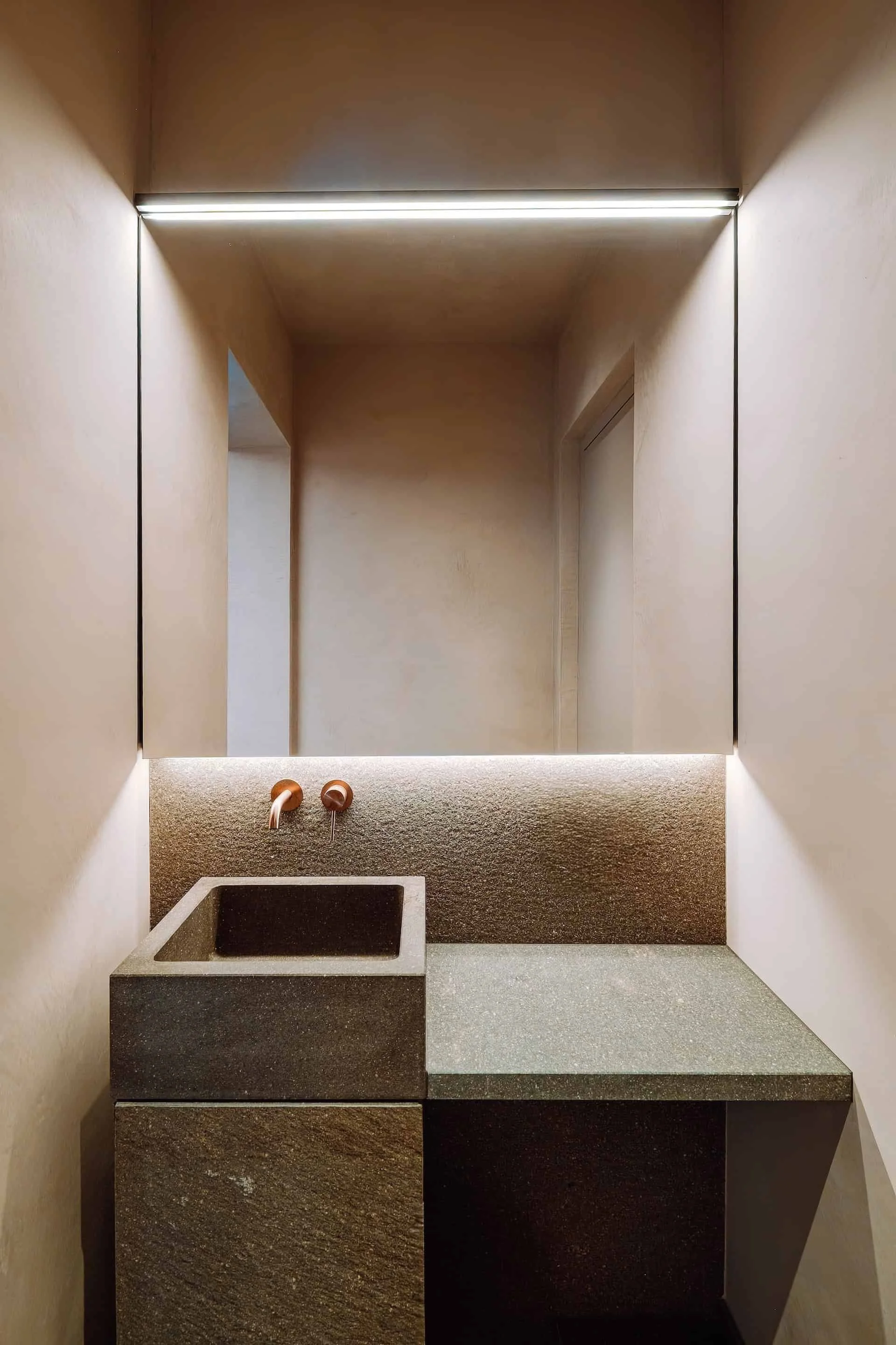 Ein moderner Waschraum mit einer kleinen Spüle aus schwarzem Granit und einem Handwaschbecken mit Kupferarmaturen. Die Wände sind in einem neutralen Sand- oder Beige-Ton gehalten, mit einer minimalistischen Beleuchtung im oberen Bereich.