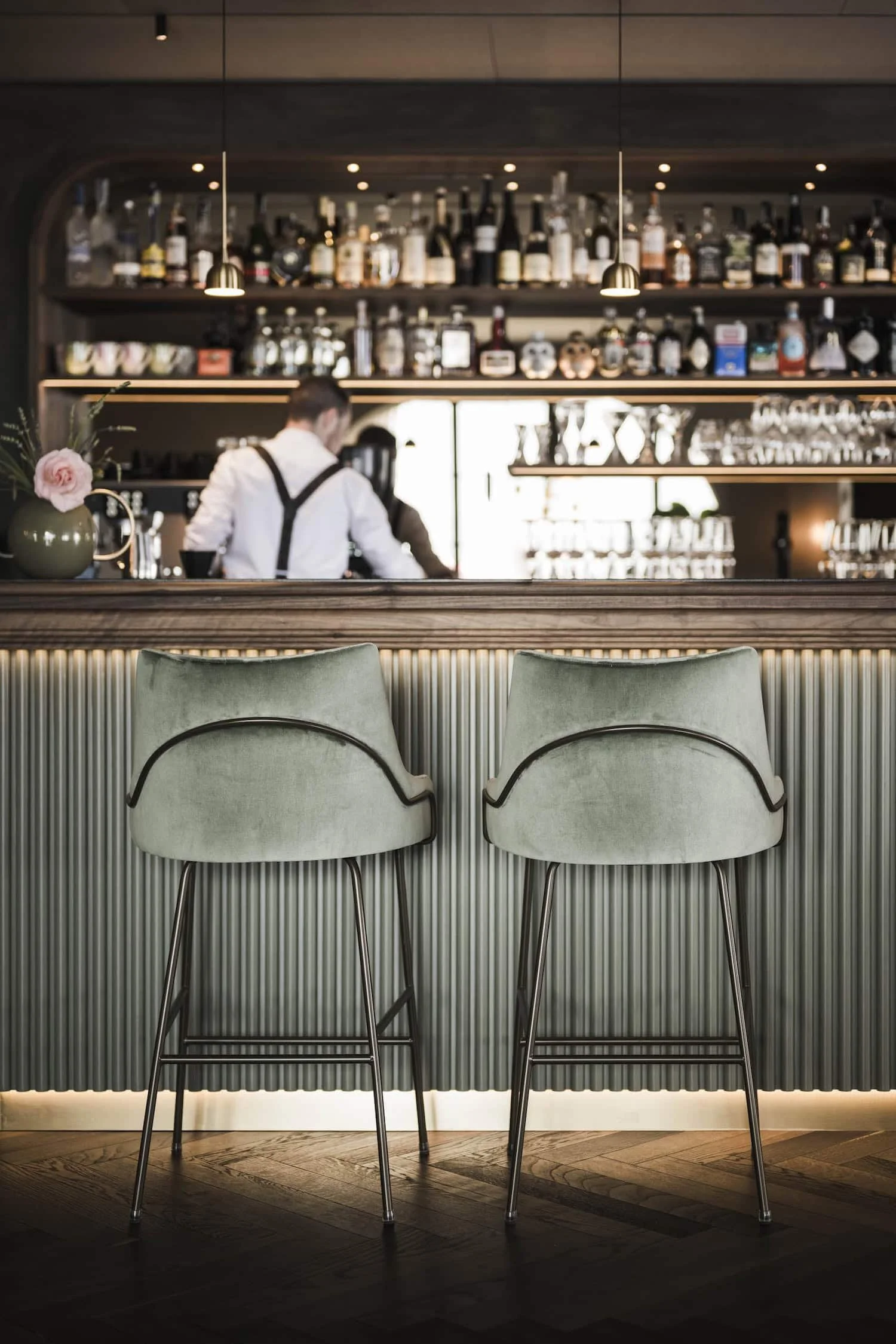Blick auf eine Bar mit hohen Stühlen, im Hintergrund Regale mit Flaschen und Gläsern, eine Vase mit Blume auf der Theke, im Vordergrund Sitzmöbel mit grünen Kissen.