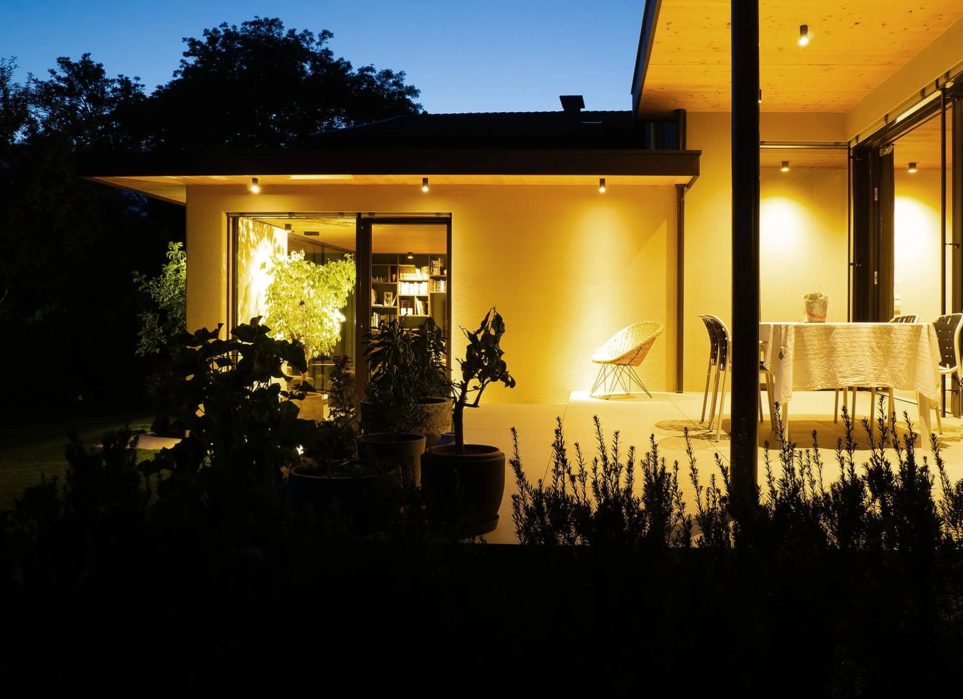 Ein modernes Haus mit blauer Dämmerung im Hintergrund, hell erleuchtete Terrasse mit Gartenpflanzen, Esstisch und Stühlen, große Fenster mit Bücherregalen im Inneren.